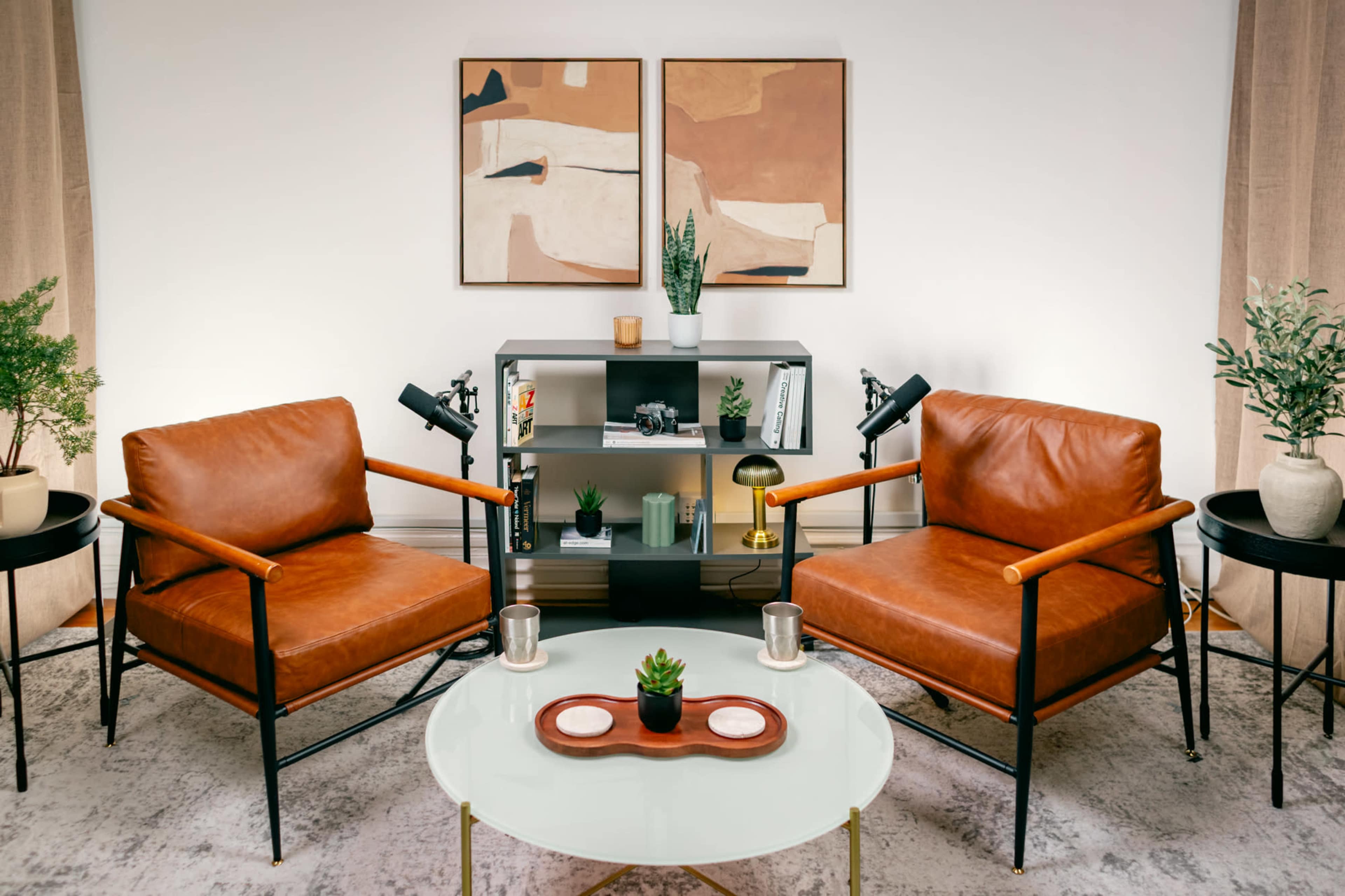 The image shows a modern living room setup with two brown leather chairs facing a small round table, accompanied by a bookshelf and decorative plants.