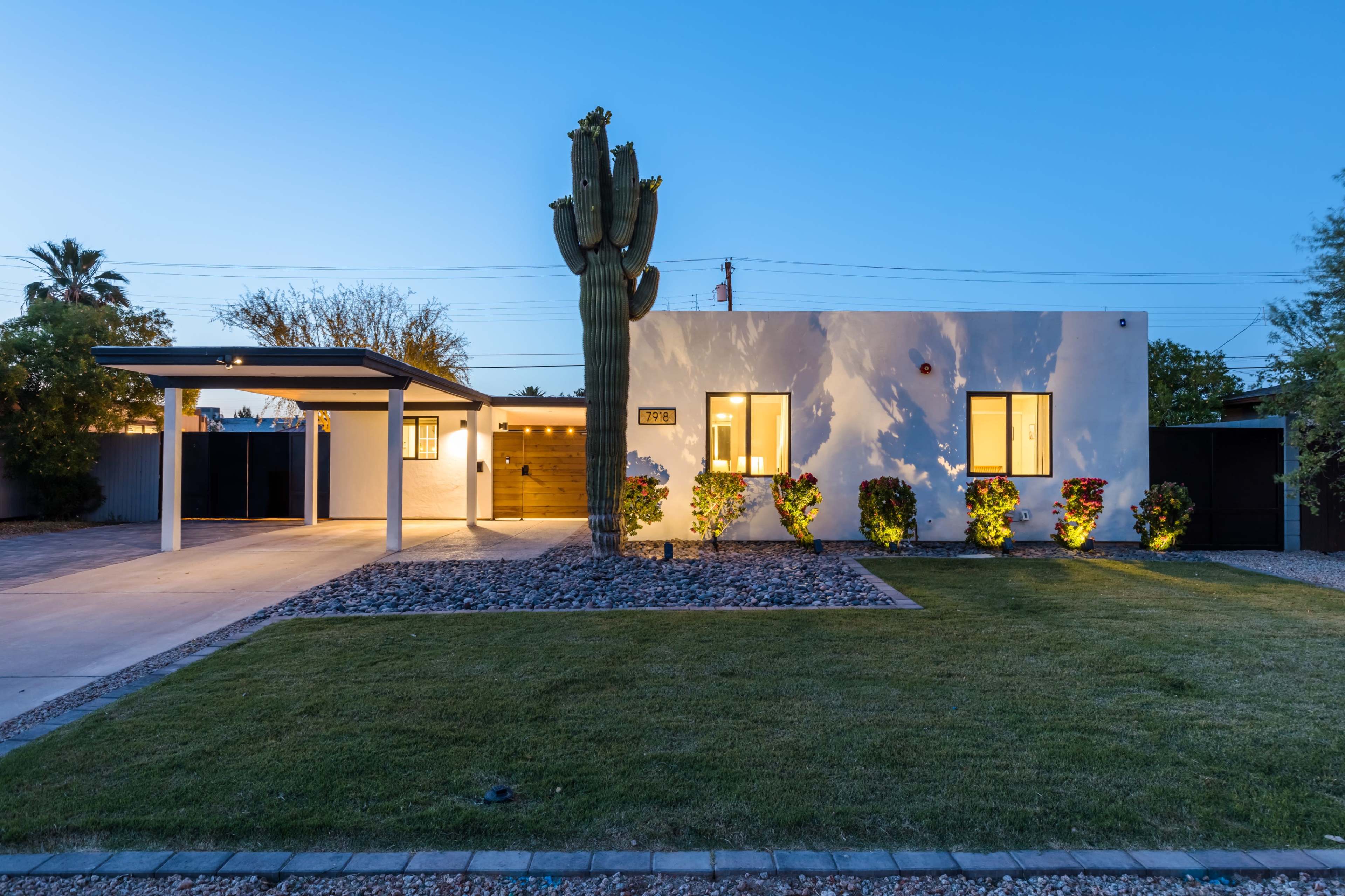 A modern white house with large windows and a carport is situated in a landscaped yard featuring a prominent cactus and gravel accents.