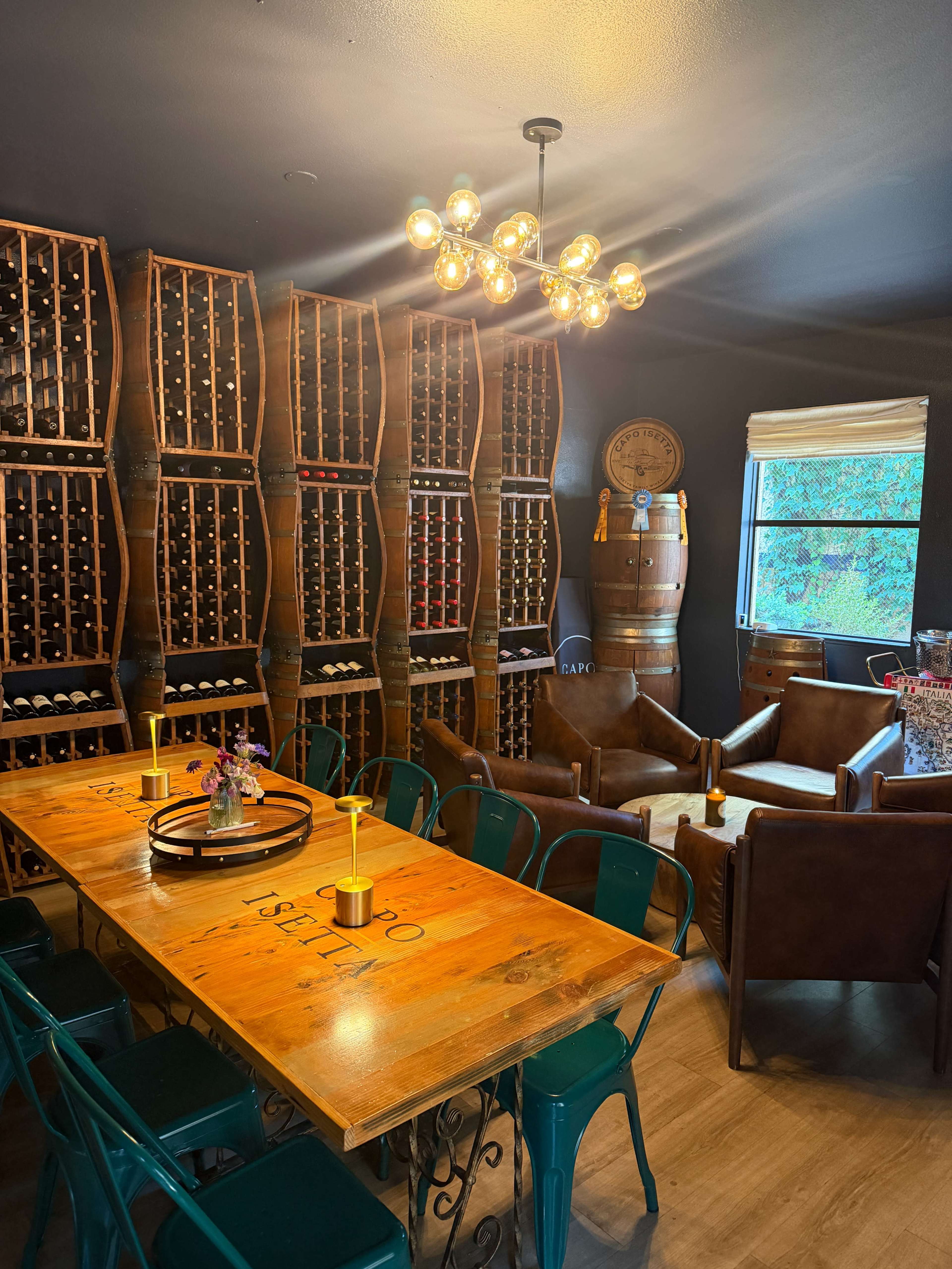 The image shows a wine cellar with wooden racks filled with bottles, a large wooden table in the center, and leather chairs arranged nearby.