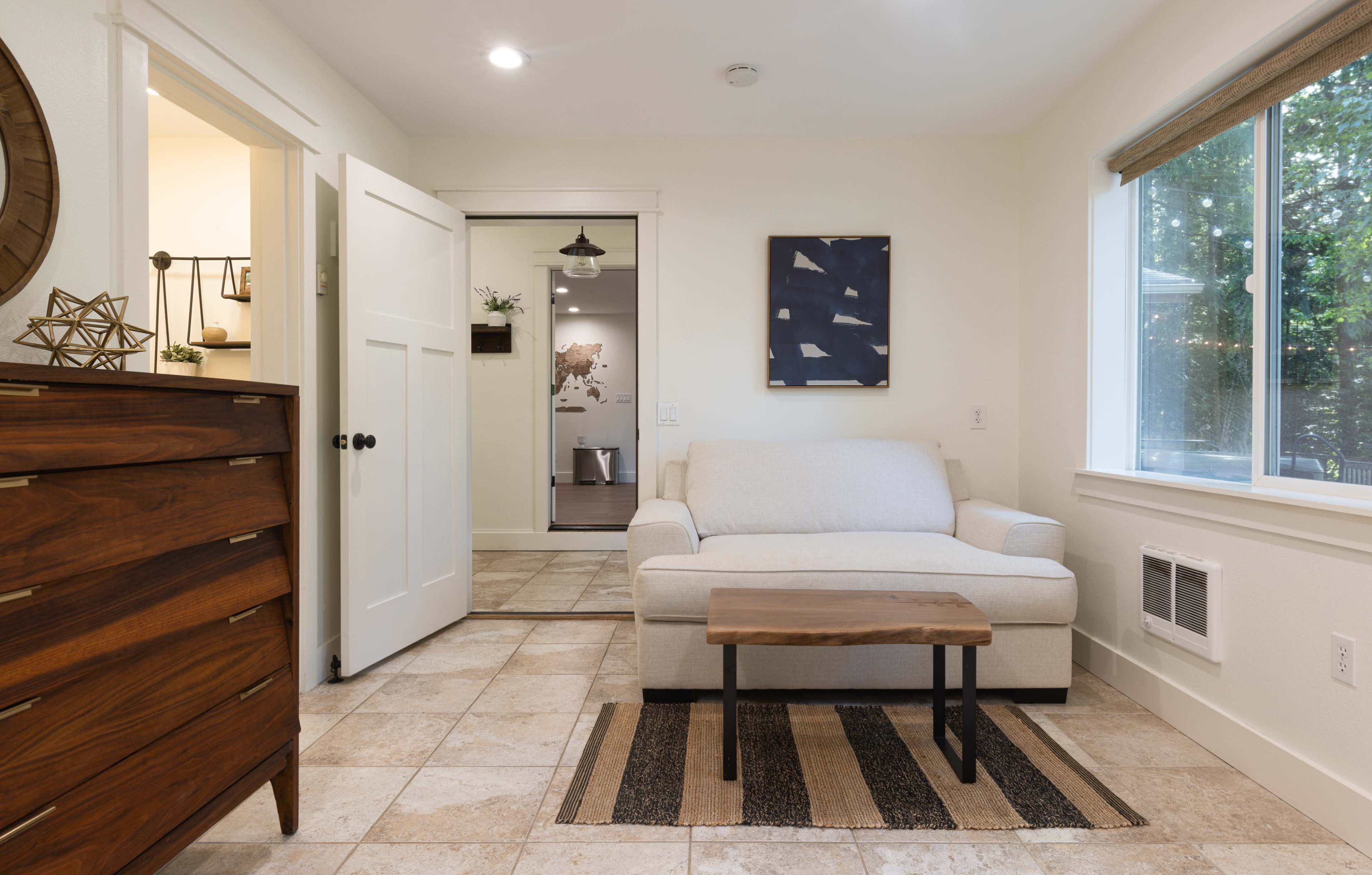 A small, well-lit room featuring a light-colored sofa, a wooden coffee table, and a chest of drawers, with a doorway leading to another area.