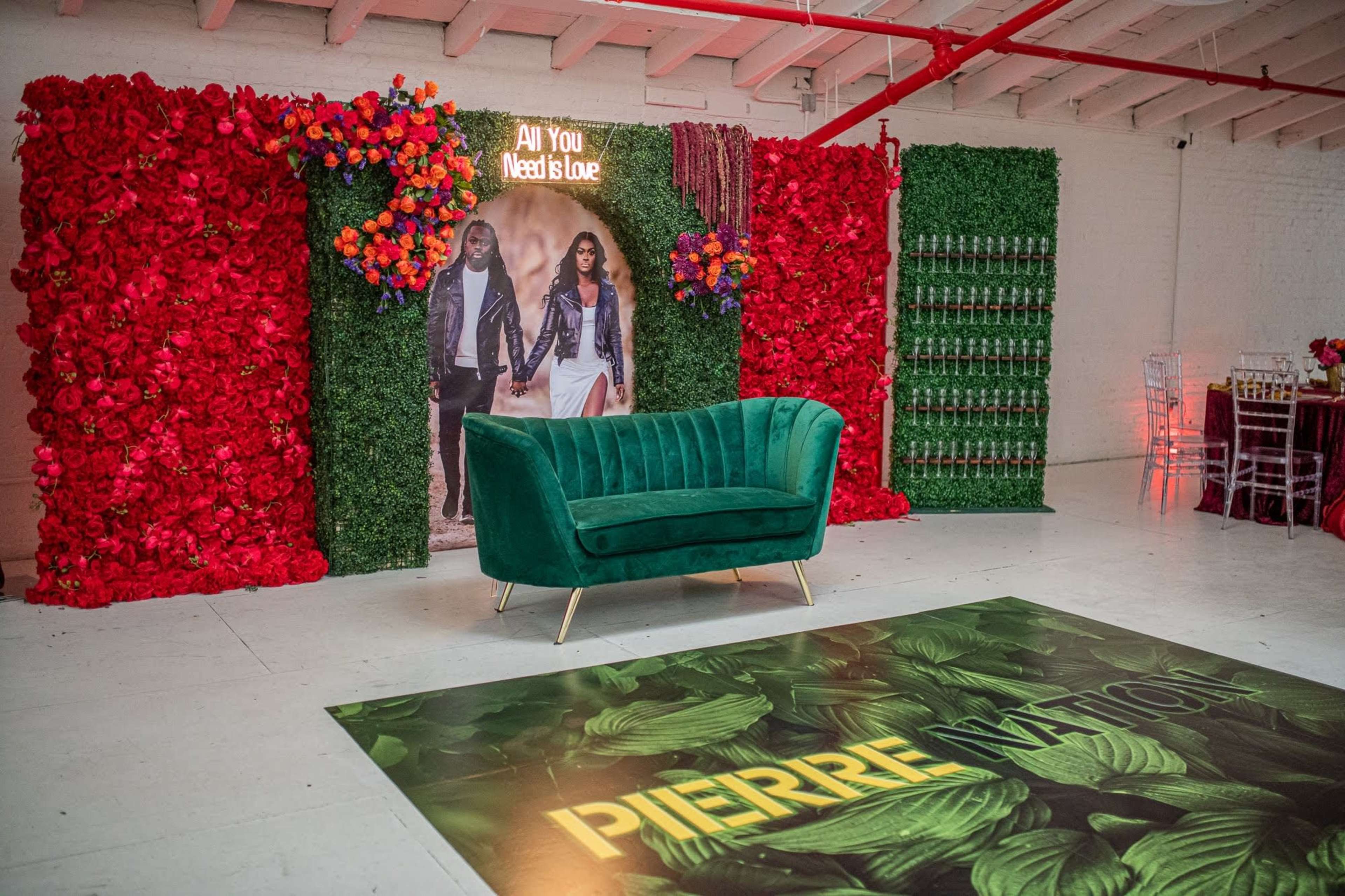 A decorated event space featuring a green velvet couch in front of a floral backdrop, a large poster, and an abundance of bottled drinks on display.