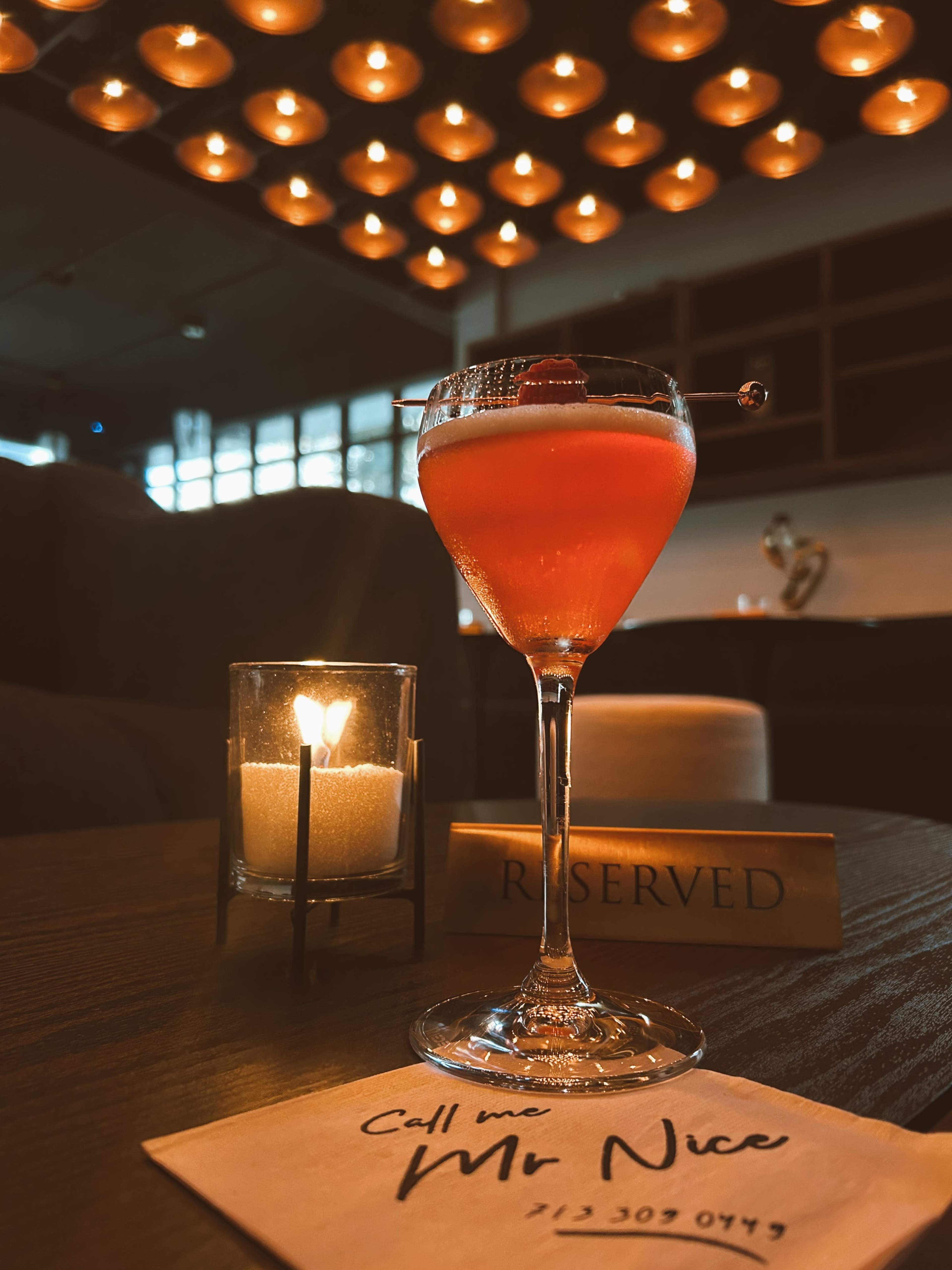 A cocktail served in a stemmed glass sits on a table next to a lit candle and a reserved sign, with a napkin that reads "Call me Mr. Nice."