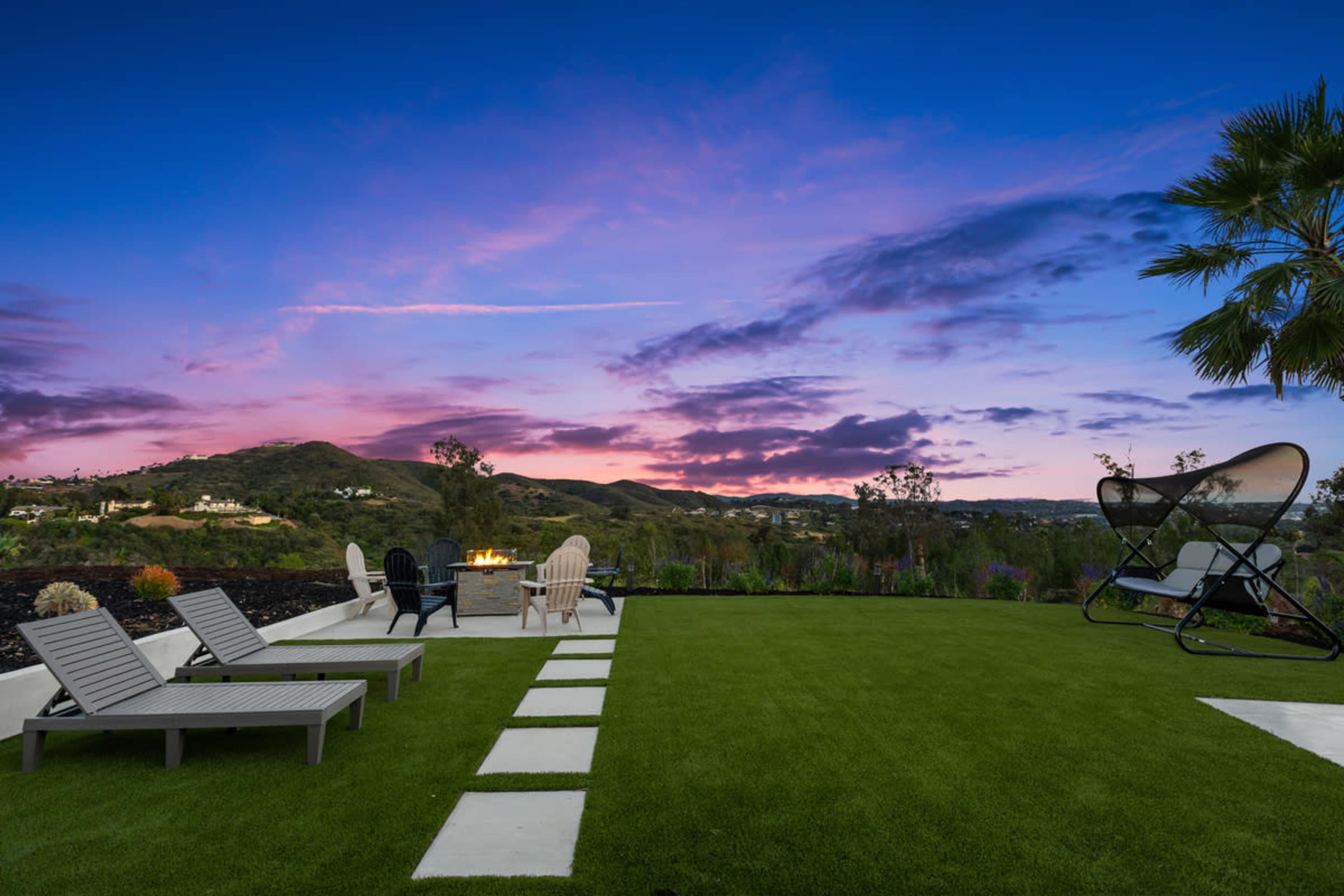 The image depicts a landscaped backyard featuring lounge chairs, a fire pit, and a scenic view of hills under a colorful sunset sky.