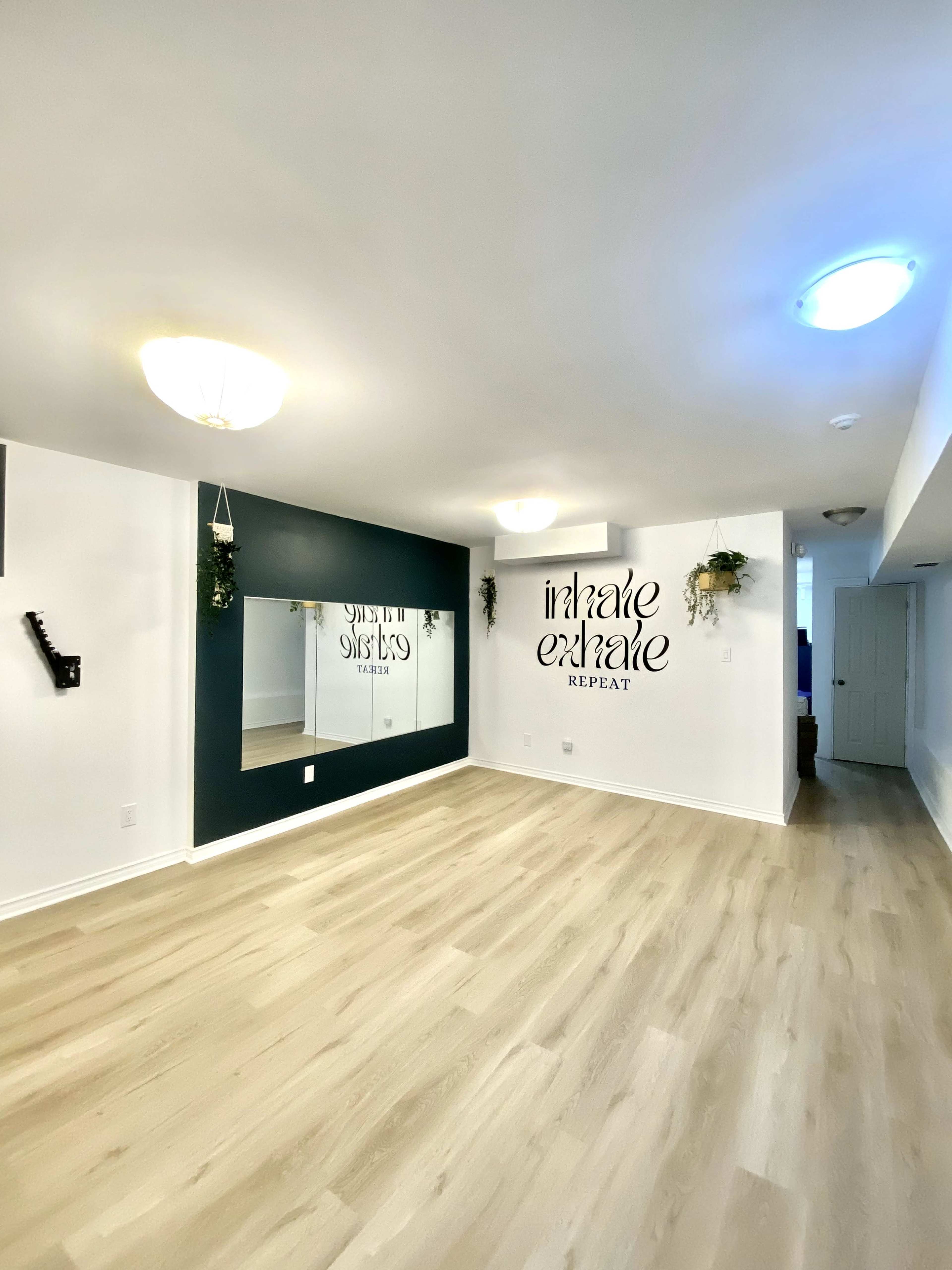 The image shows a spacious room with light wood flooring, a large mirror on one wall, and text that reads "inhale exhale repeat" on another wall.