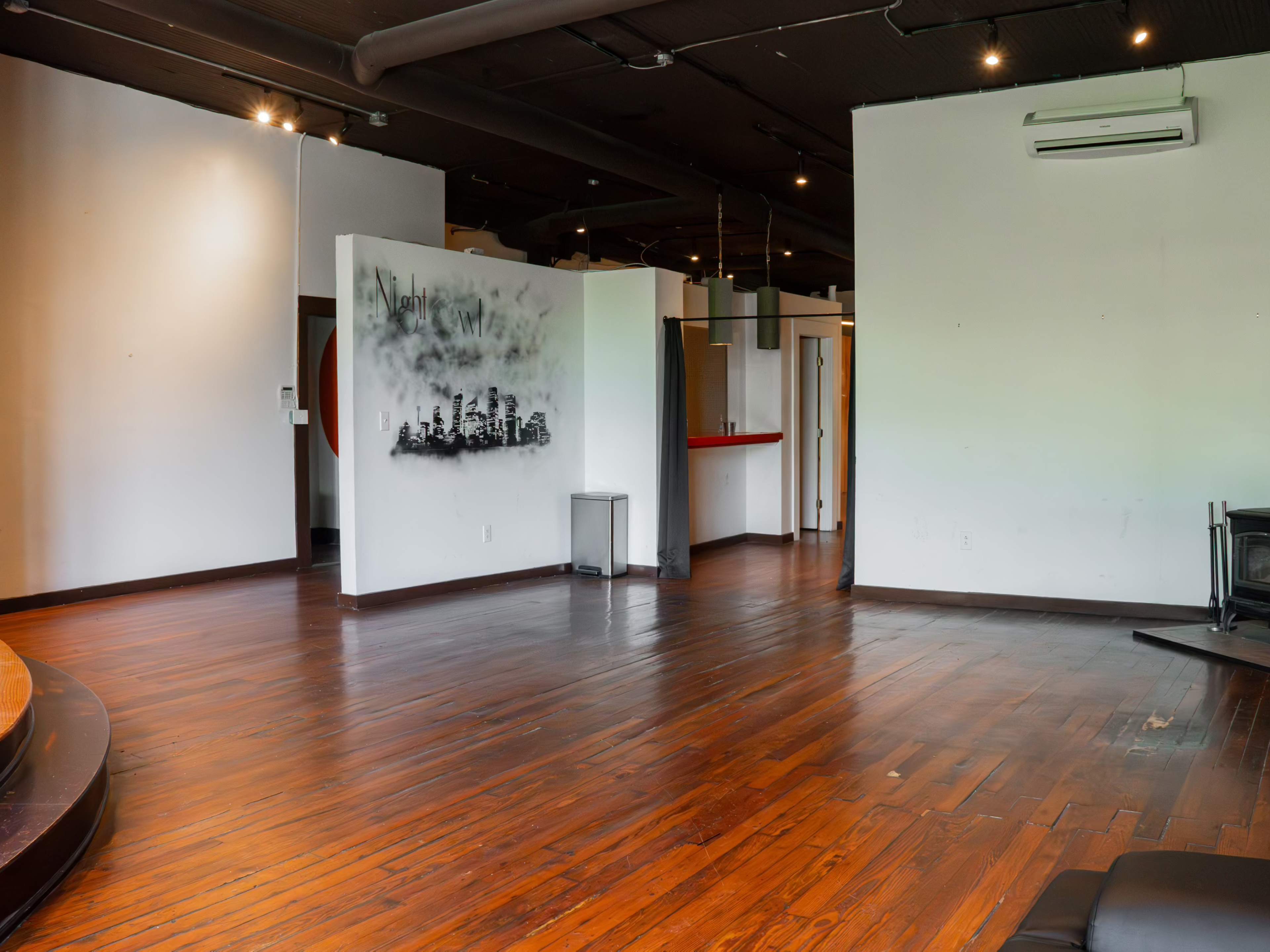 An open room with wooden flooring, a wall featuring a city skyline mural, and several areas including a kitchenette and a small partitioned space.