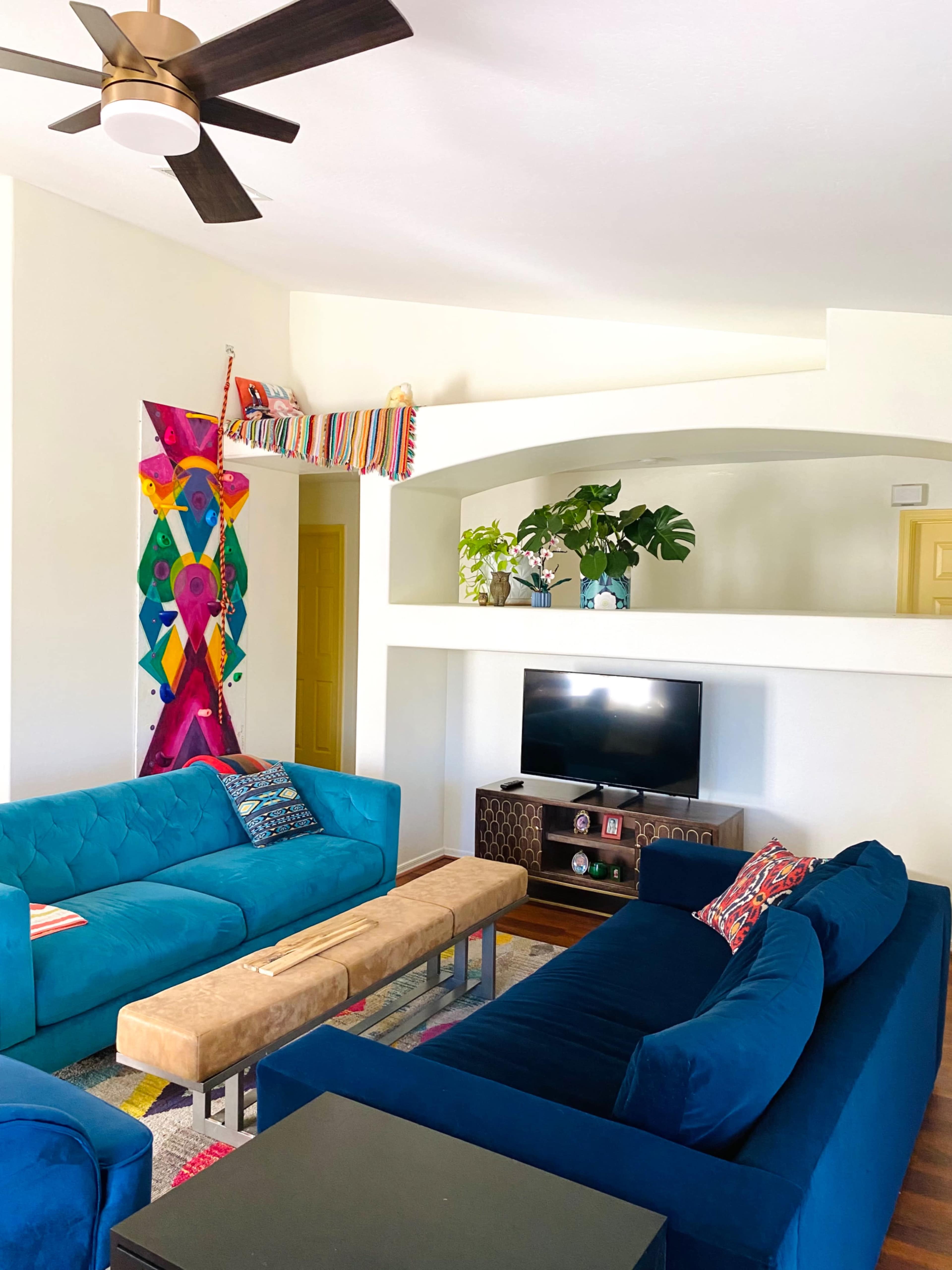 A living room features blue sofas, a wooden coffee table, a television stand, and a colorful wall mural with plants displayed on shelves.