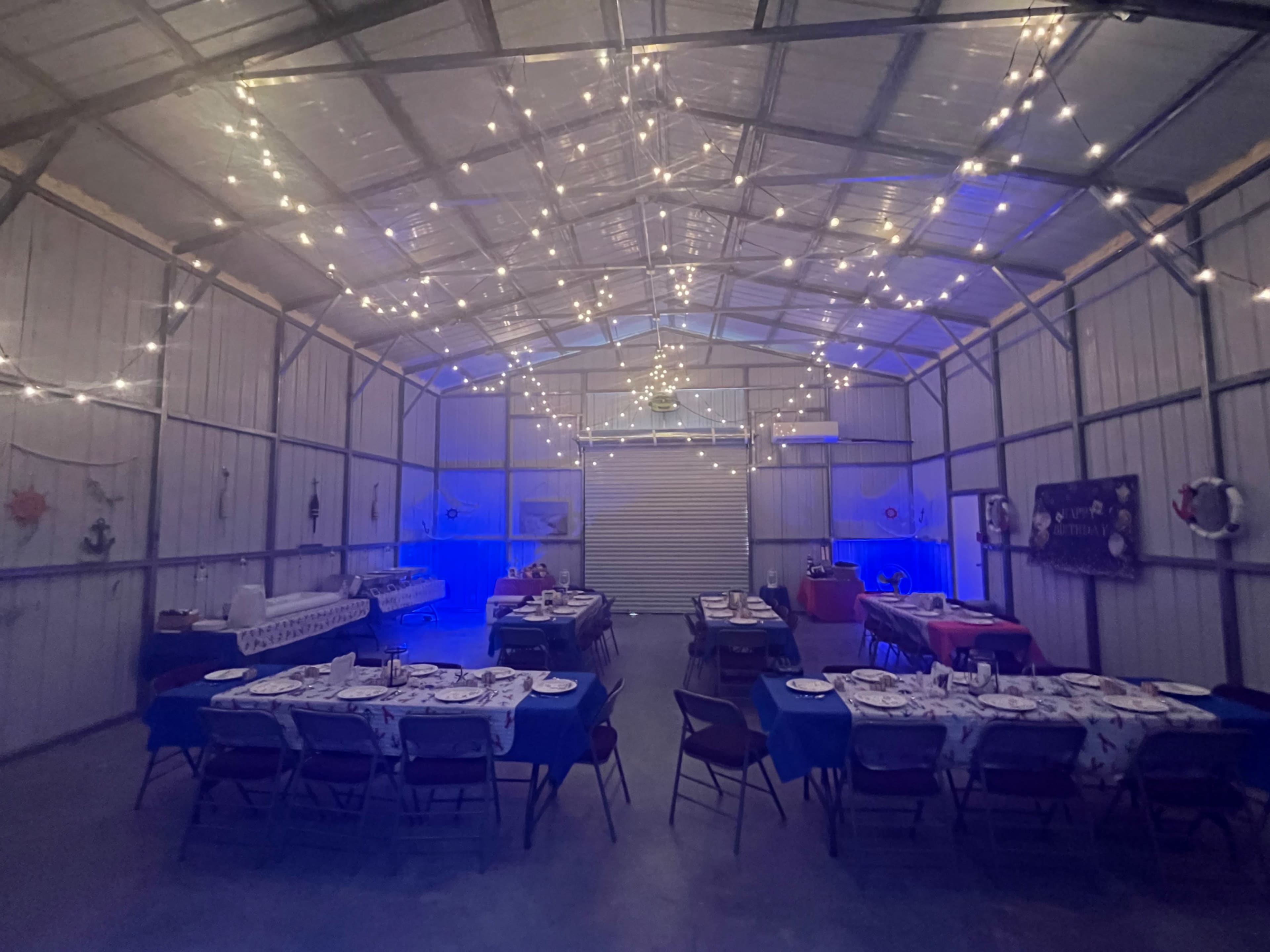 A spacious indoor venue is set up for a gathering, featuring decorated tables with place settings under string lights illuminating the metal structure.