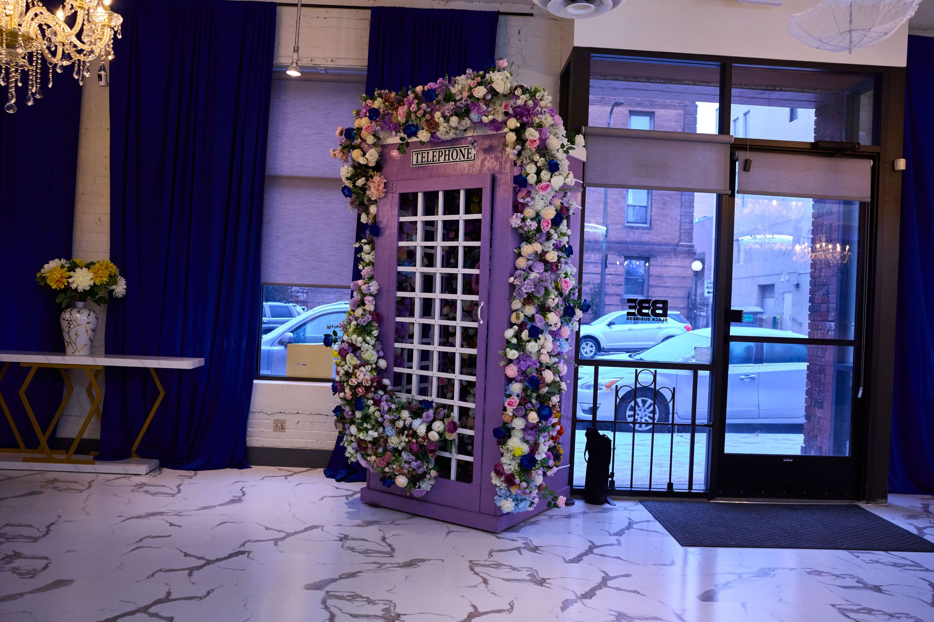 A large, purple phone booth decorated with artificial flowers stands against a wall in a brightly lit interior space.