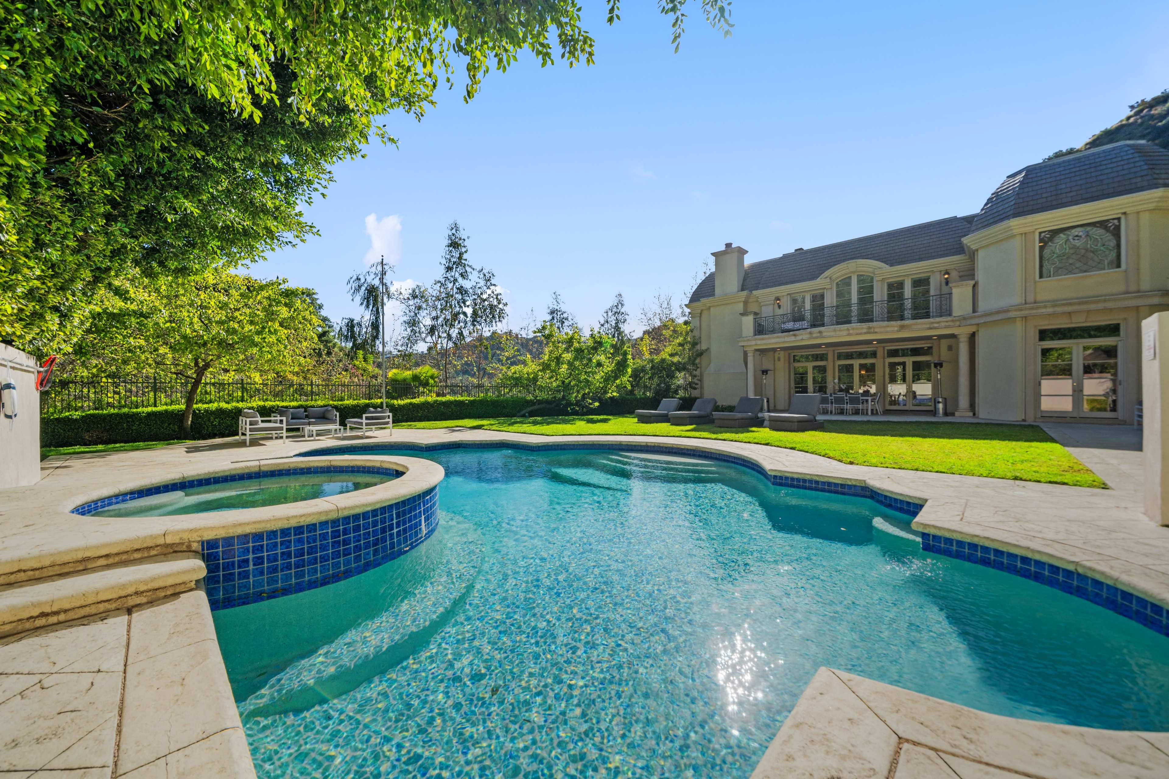 A spacious backyard features a swimming pool with a spa area, surrounded by green lawn and trees, next to a large, two-story house.