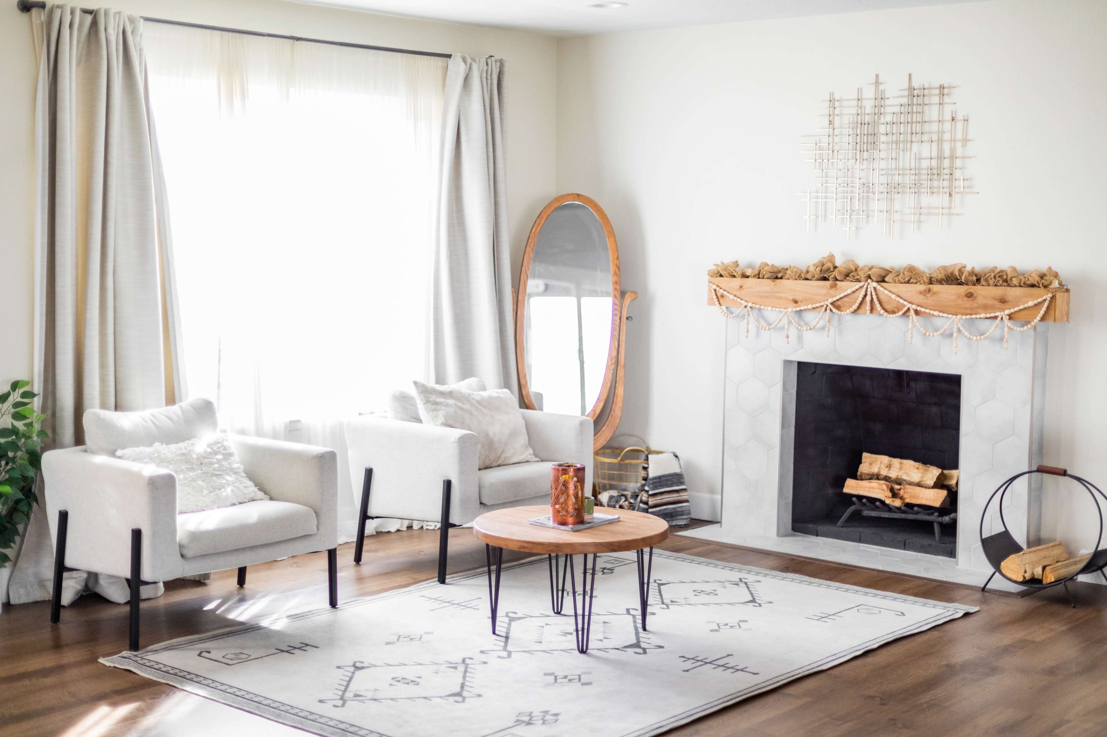 The room features two white armchairs, a round wooden coffee table, a large mirror, and a fireplace adorned with a decorative mantle and wood logs.