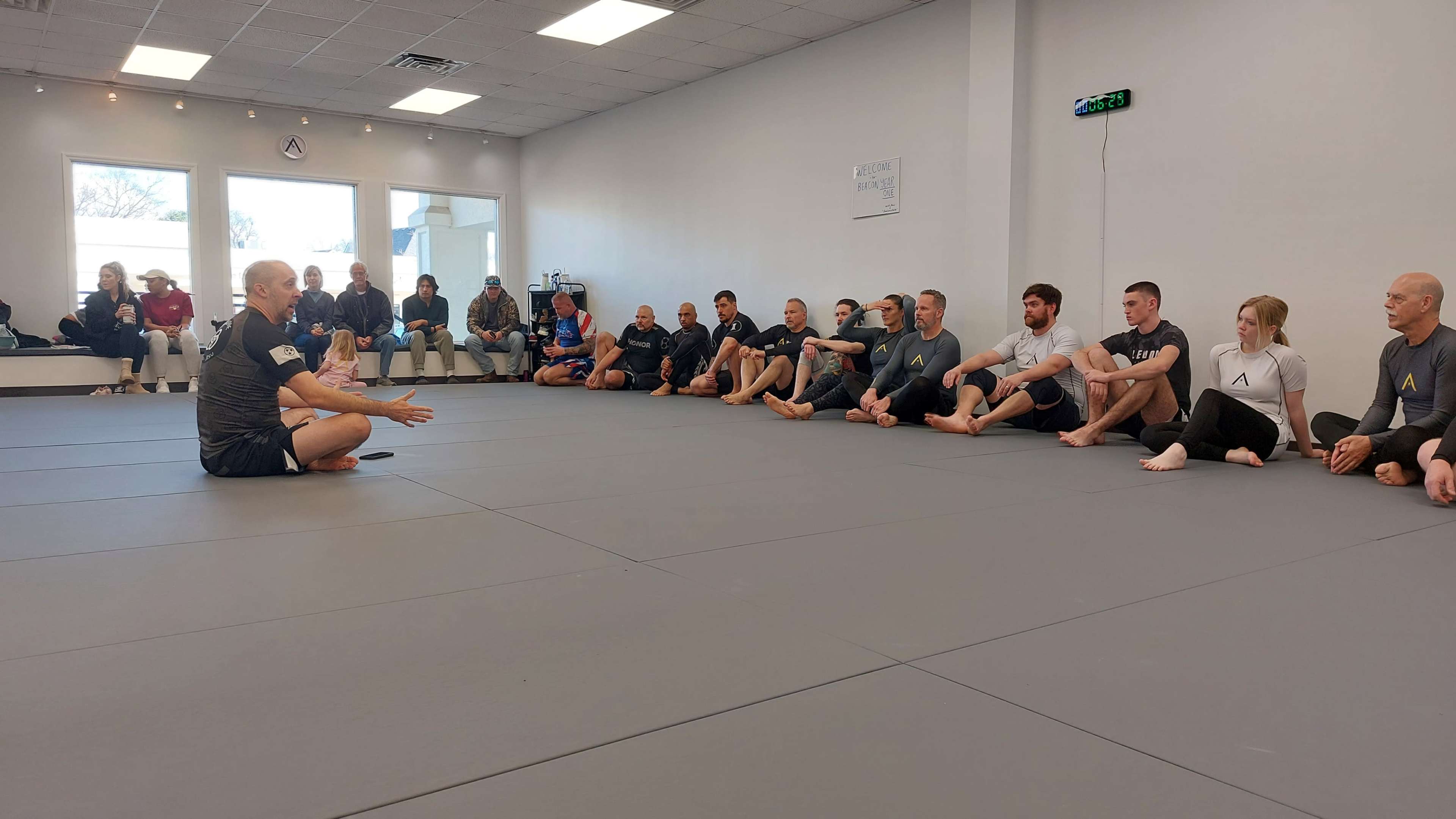 A group of individuals sits in a circle on mats, attentively listening to a person speaking in a training facility.