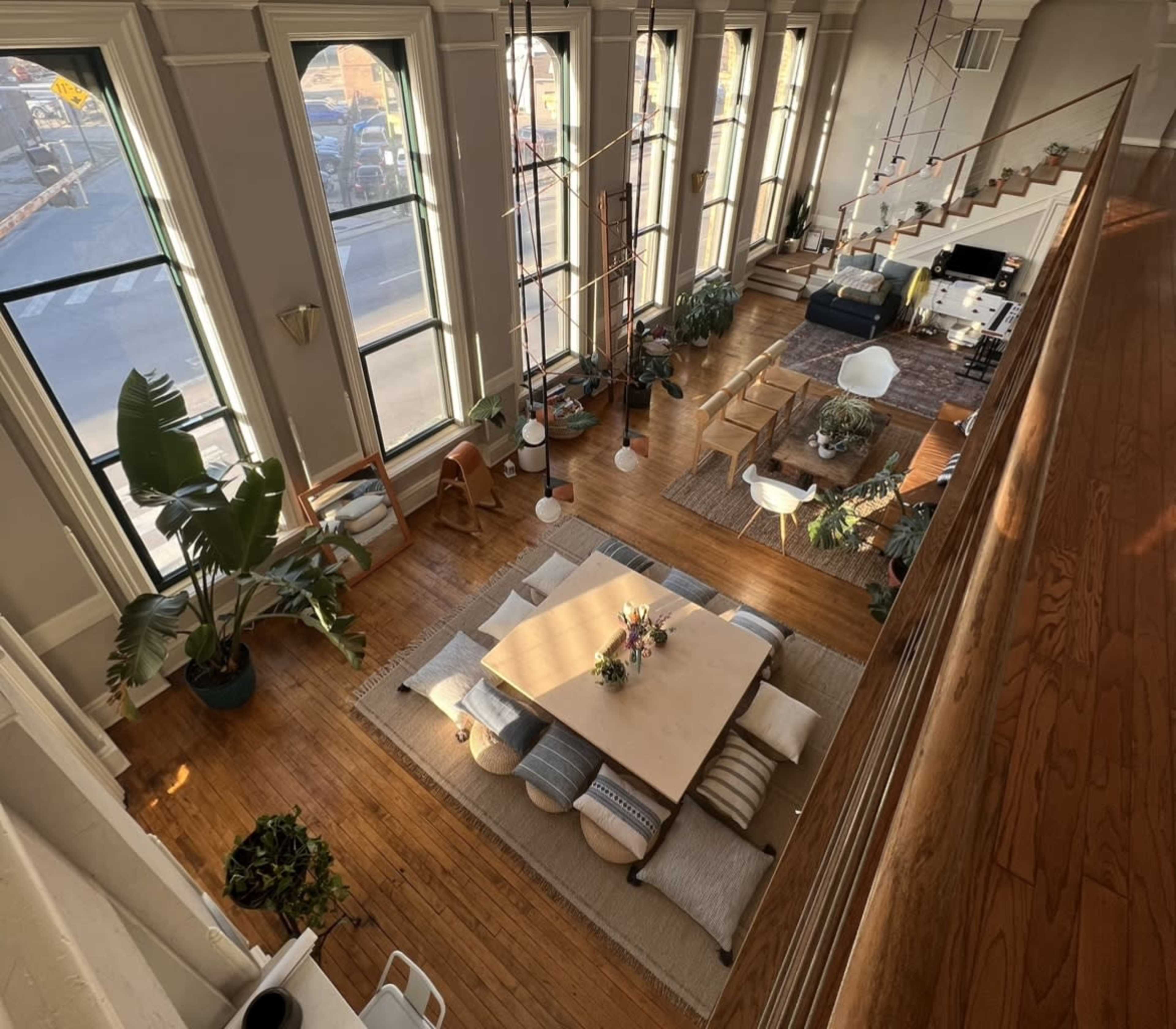 A spacious, well-lit living area with large windows, wooden flooring, and various plants, furniture arranged around a central table.