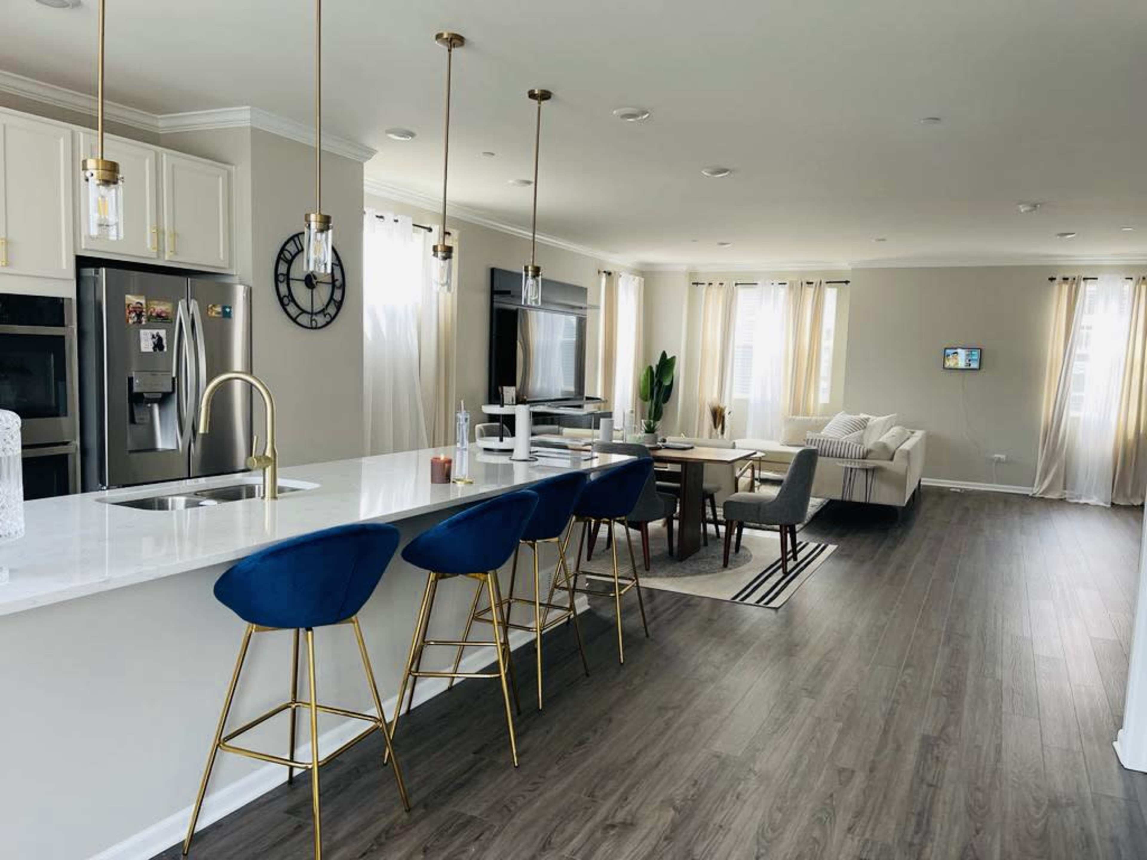 The image shows a modern kitchen and living area with a white countertop, blue bar stools, and light-colored furniture.