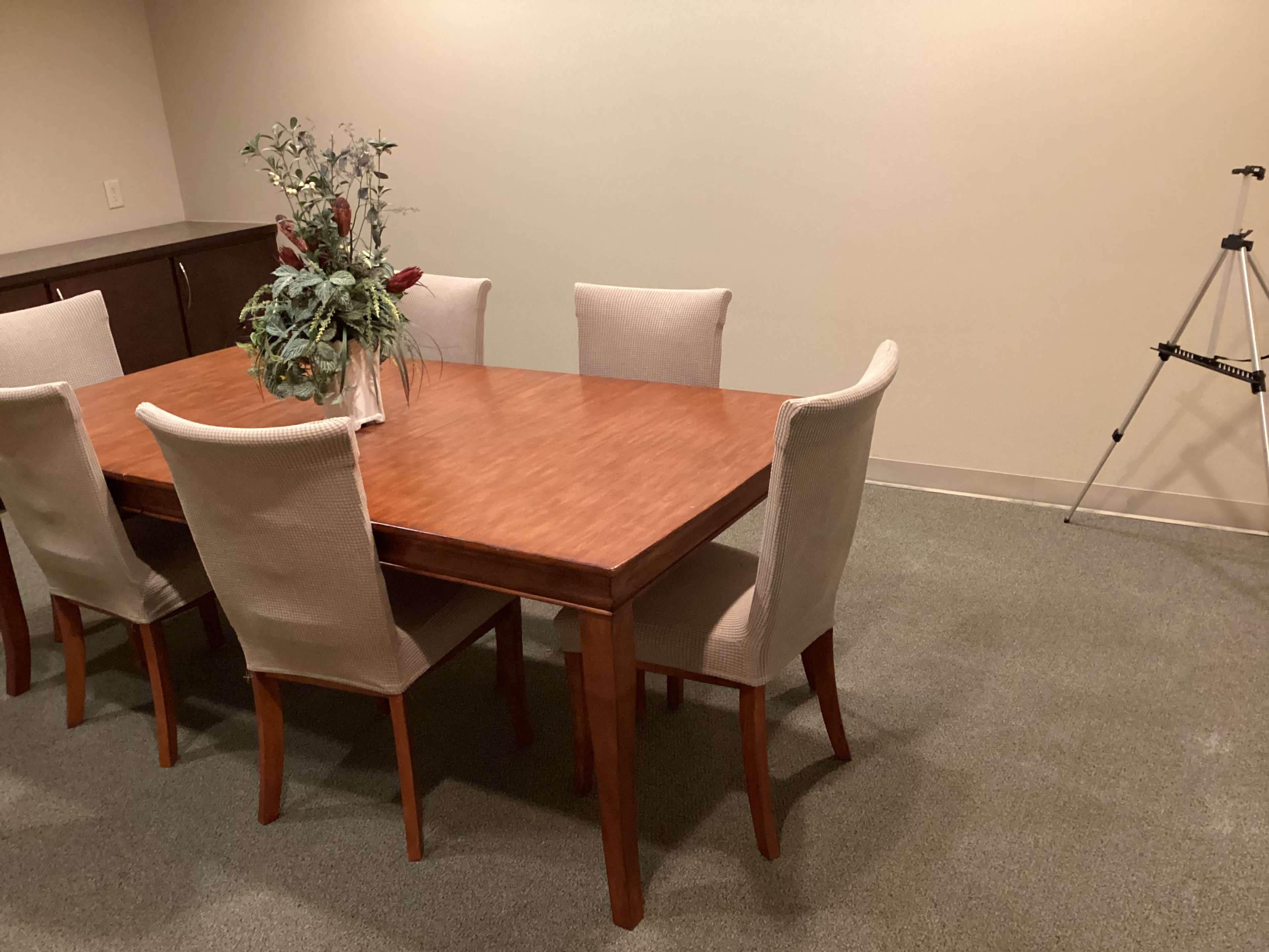 A wooden dining table surrounded by six upholstered chairs sits in a room, with a small vase of flowers on the table and a tripod positioned nearby.