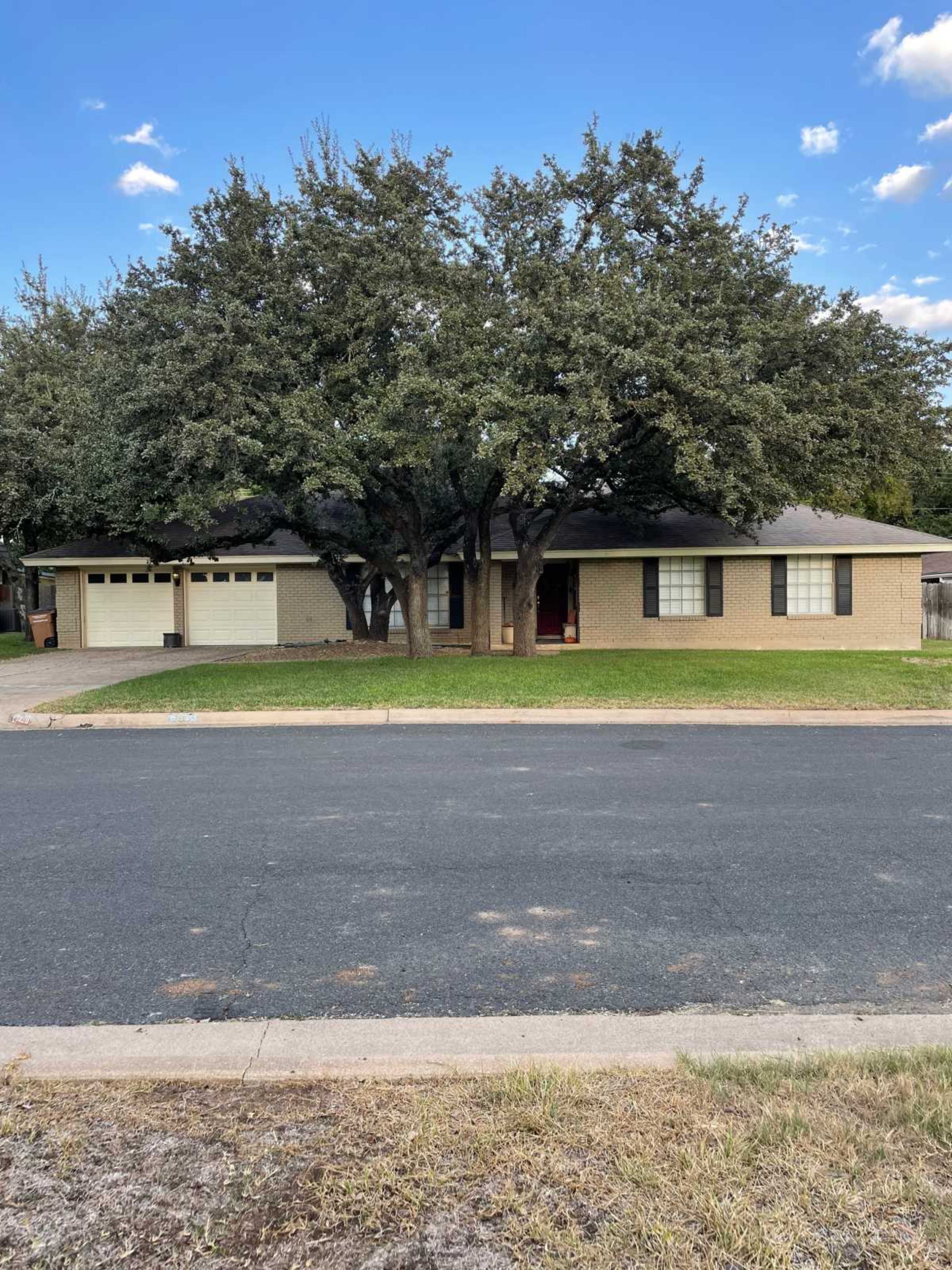 The image shows a single-story brick house with a two-car garage, situated on a grassy lawn and framed by two large trees.