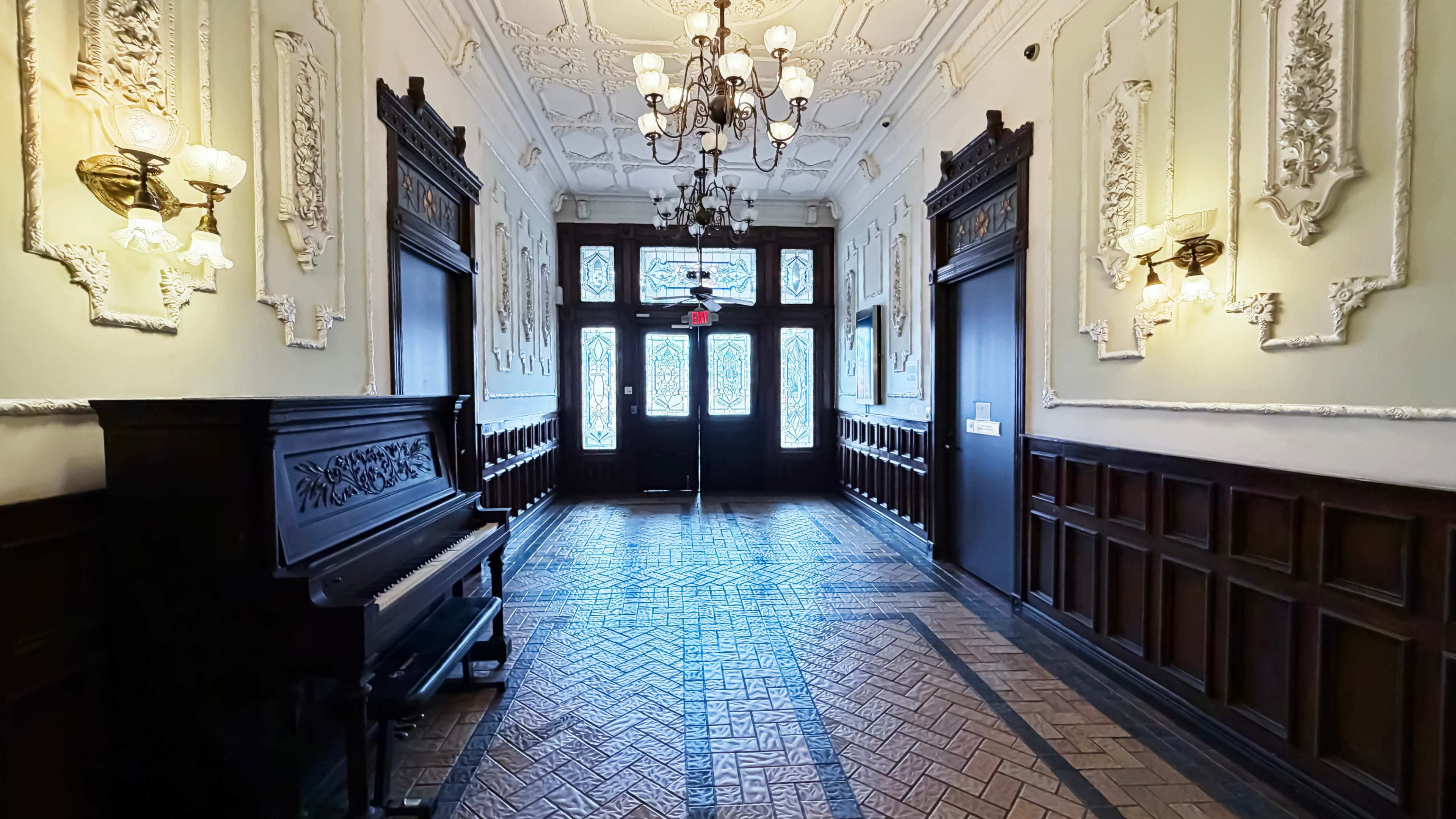 The image features a narrow hallway with ornate wall decorations, a piano on the left, and large windows at the end allowing natural light to stream in.