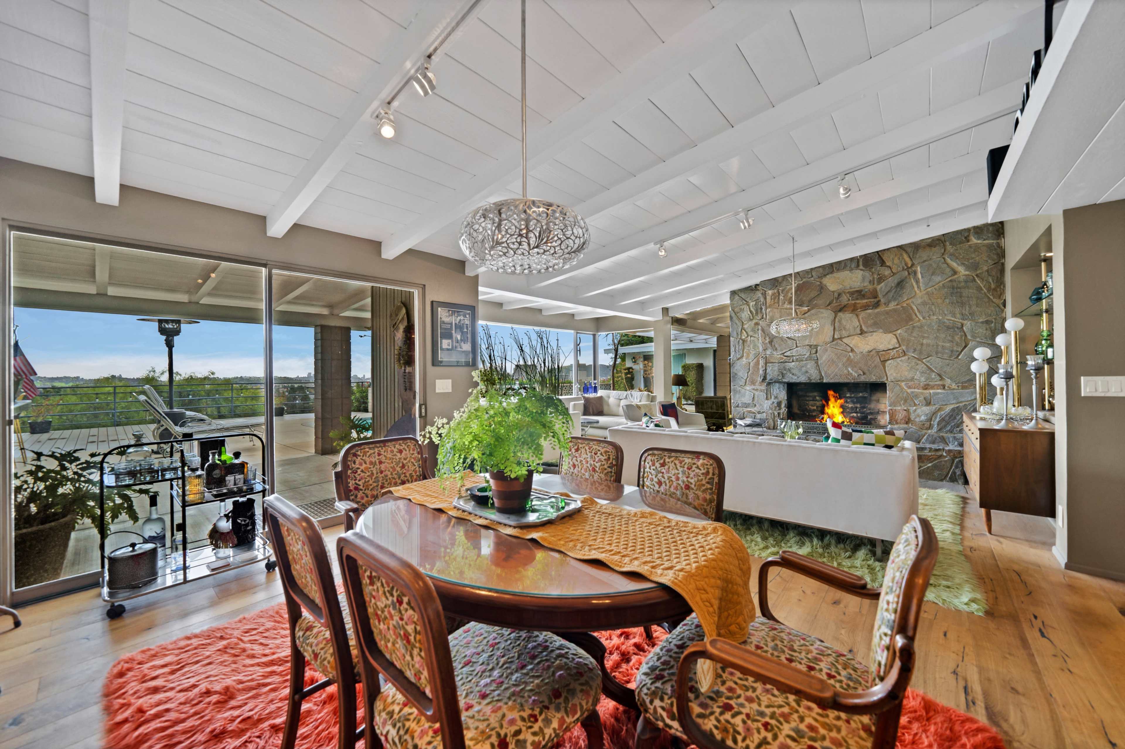 The image shows a spacious living area with a stone fireplace, a dining table set for six, and large glass doors leading to an outdoor view.