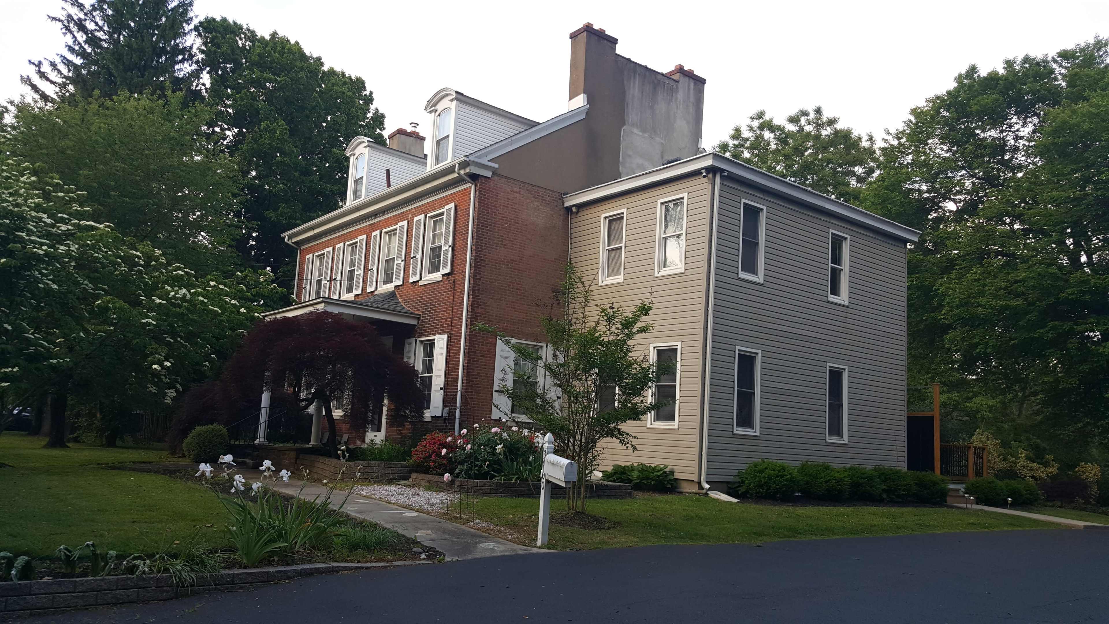 A two-story house features a mix of brick and siding with numerous windows, landscaped gardens, and a paved driveway.