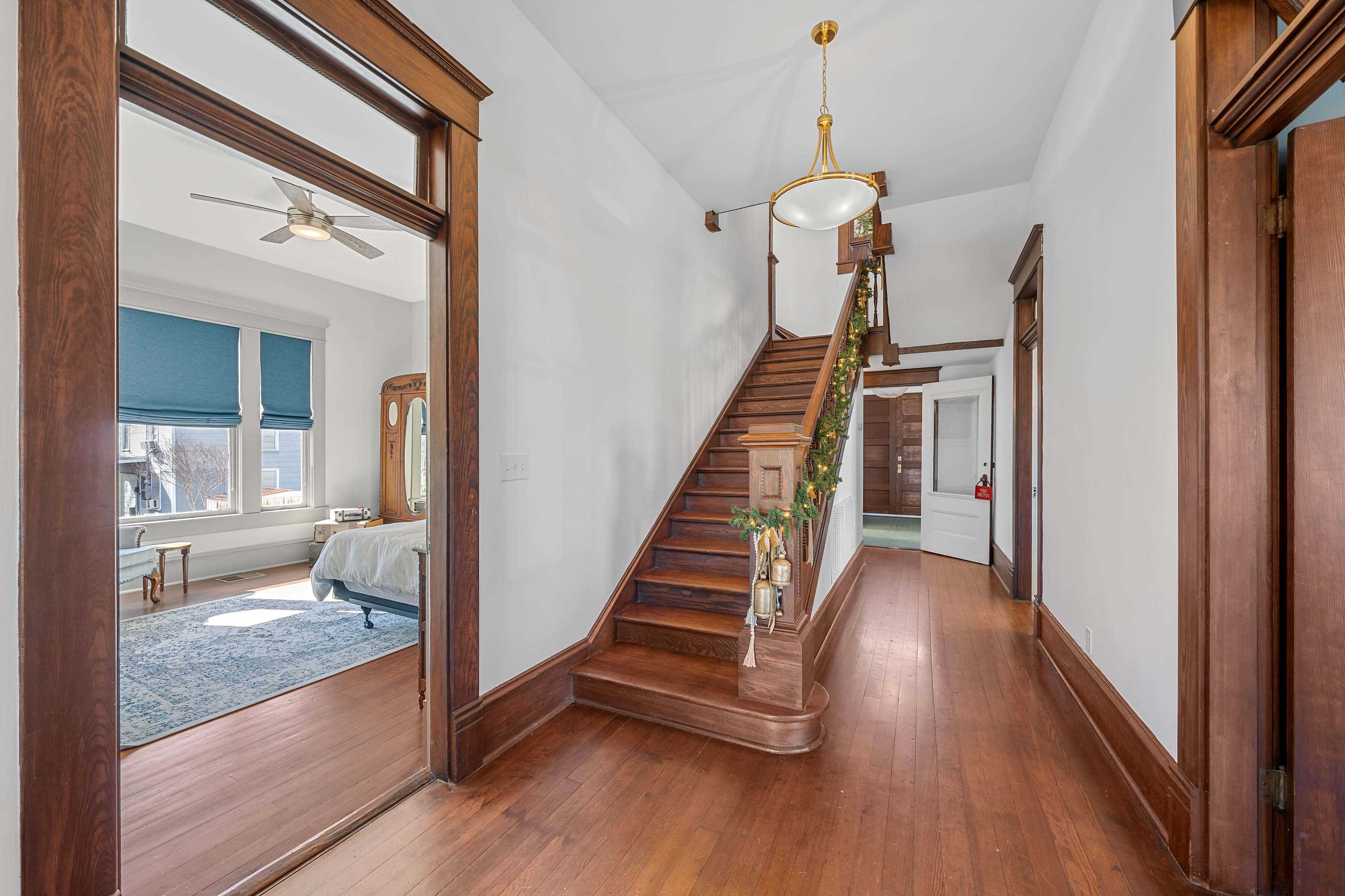 A wooden staircase in a hallway that leads to a room with windows dressed in blue shades, surrounded by wooden trim and a mirror.