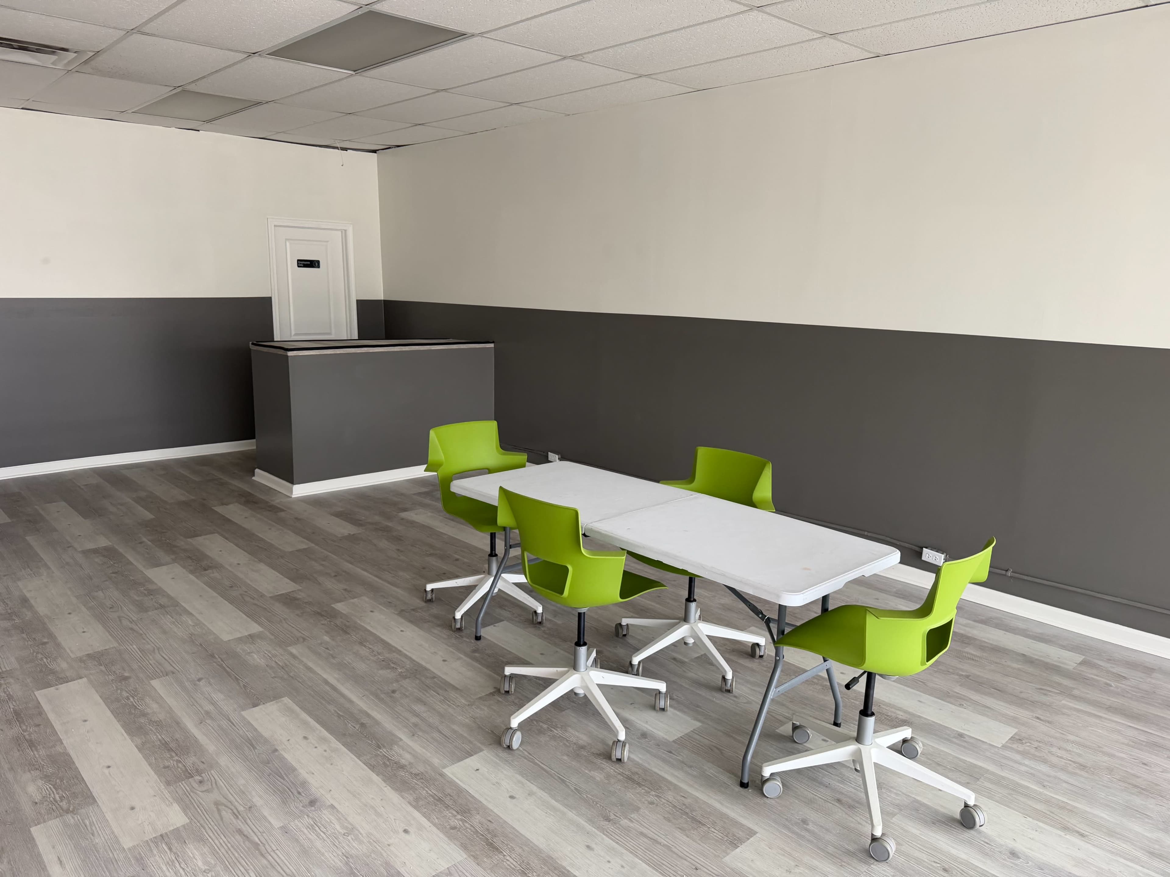 The image shows a minimalist room featuring a table and four green chairs, with a service counter and gray walls.