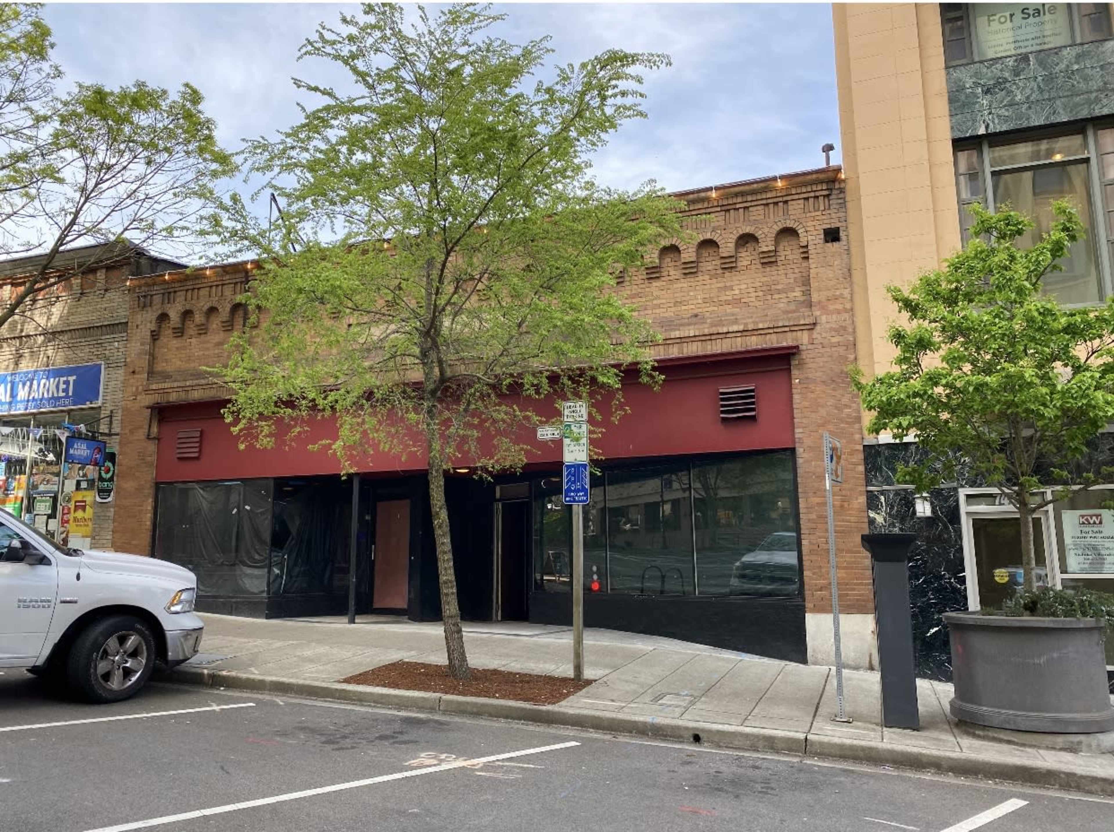 A two-story brick building with a red awning and large black windows, flanked by trees on either side.