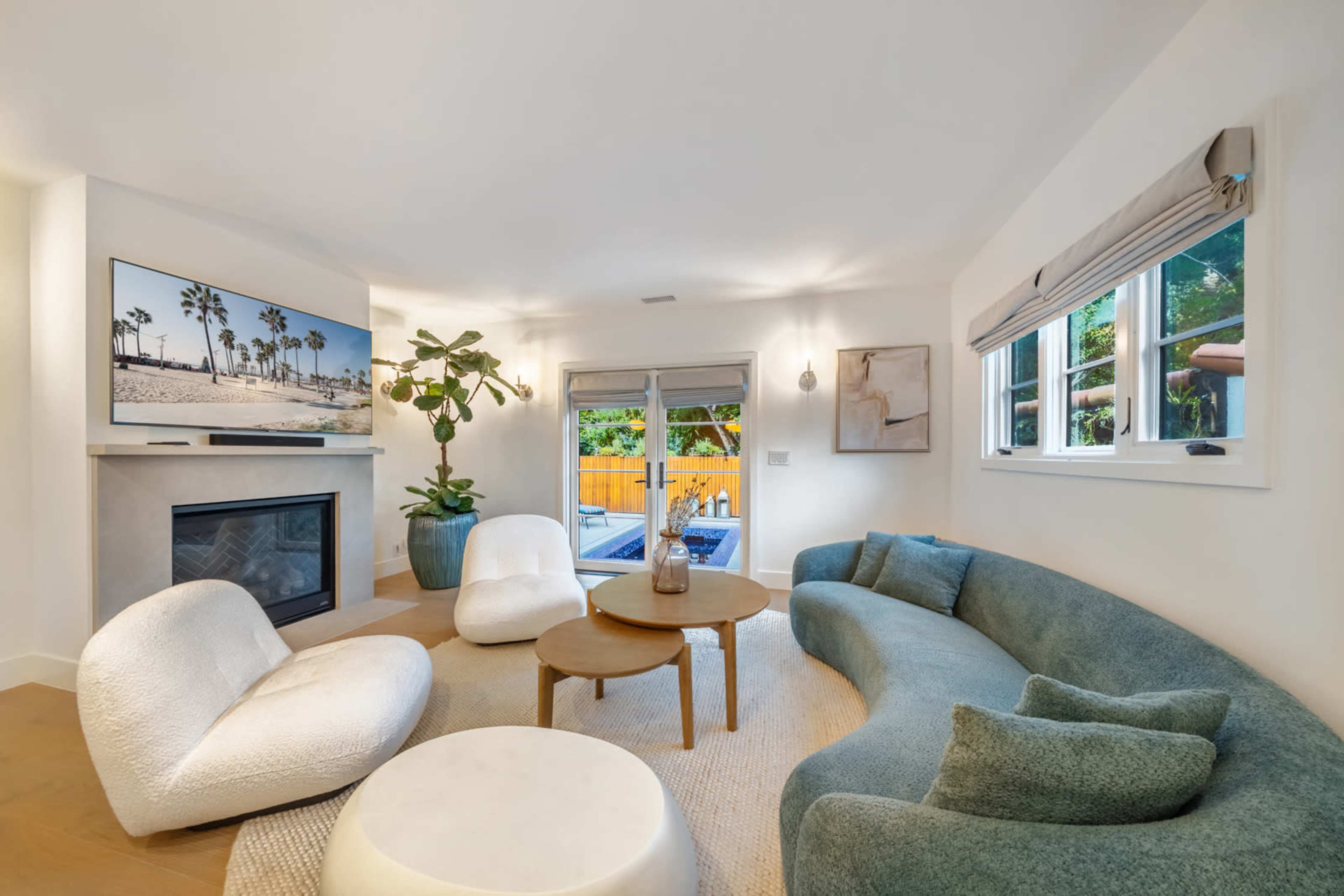 The image shows a modern living room with a curved sofa, two white armchairs, a coffee table, a fireplace, and a large television mounted on the wall.