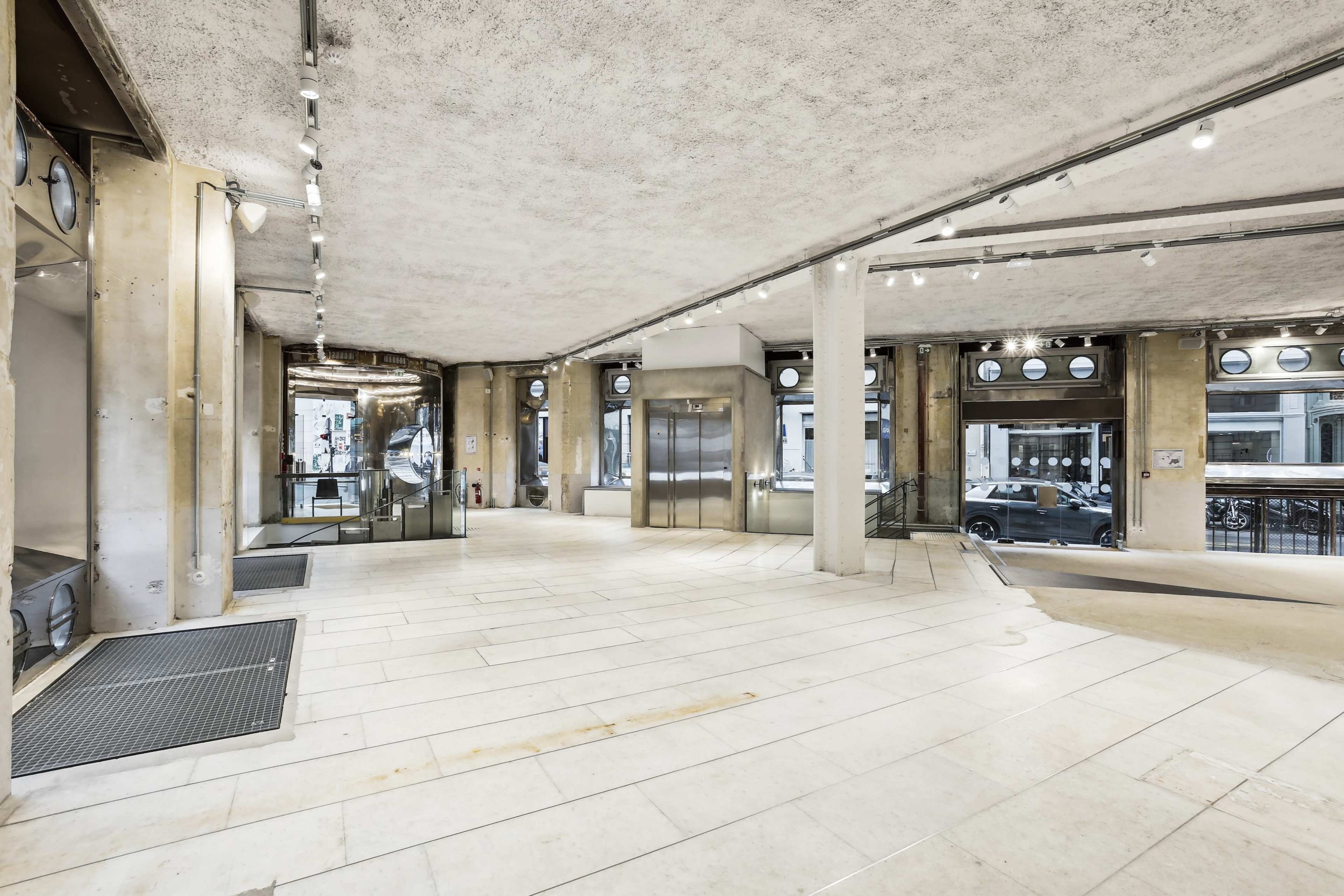 The image shows a spacious, modern interior with concrete walls and a polished floor, featuring elevators and large windows.