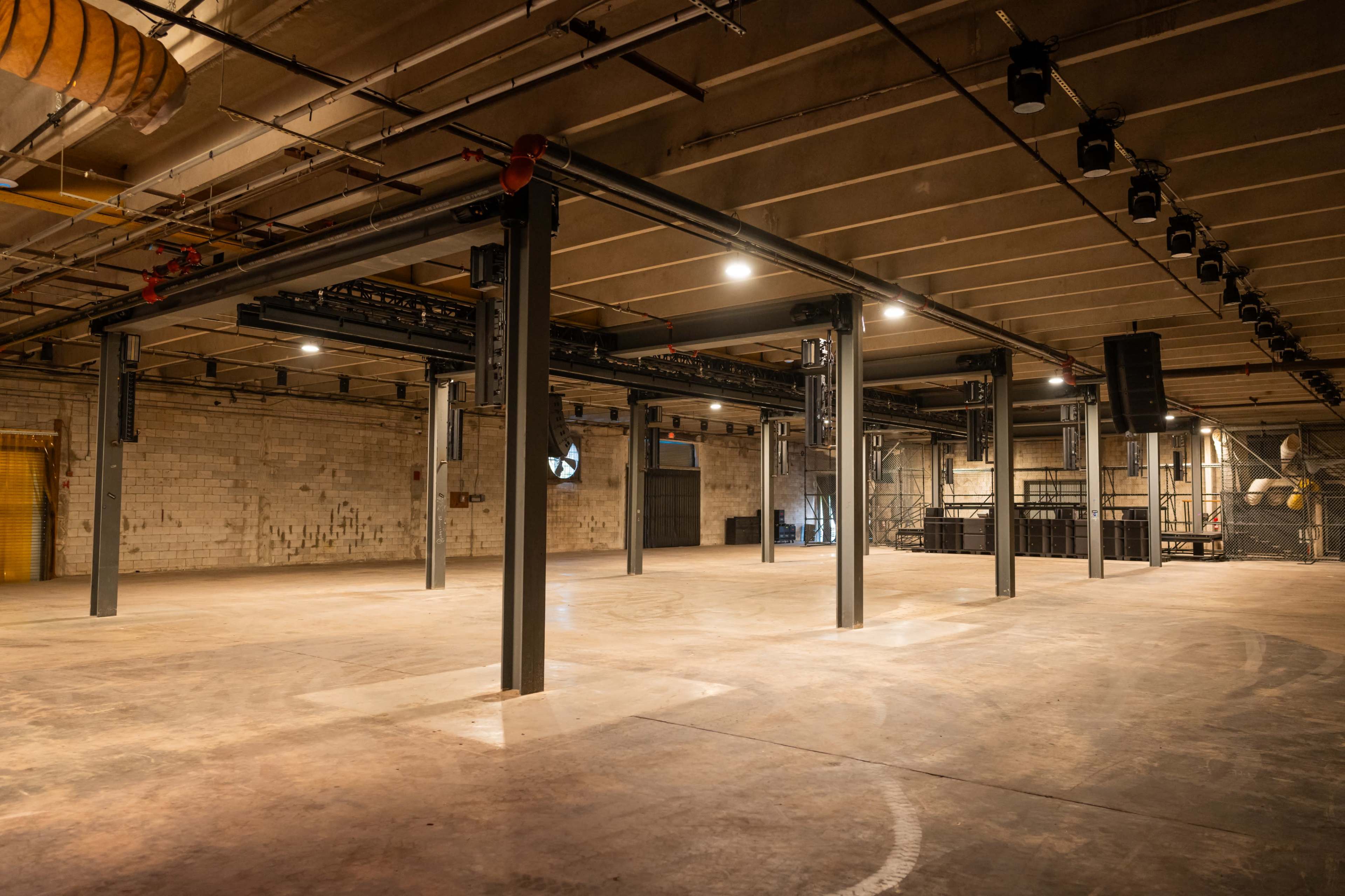The image shows an empty industrial space with exposed beams and a concrete floor, illuminated by overhead lights.