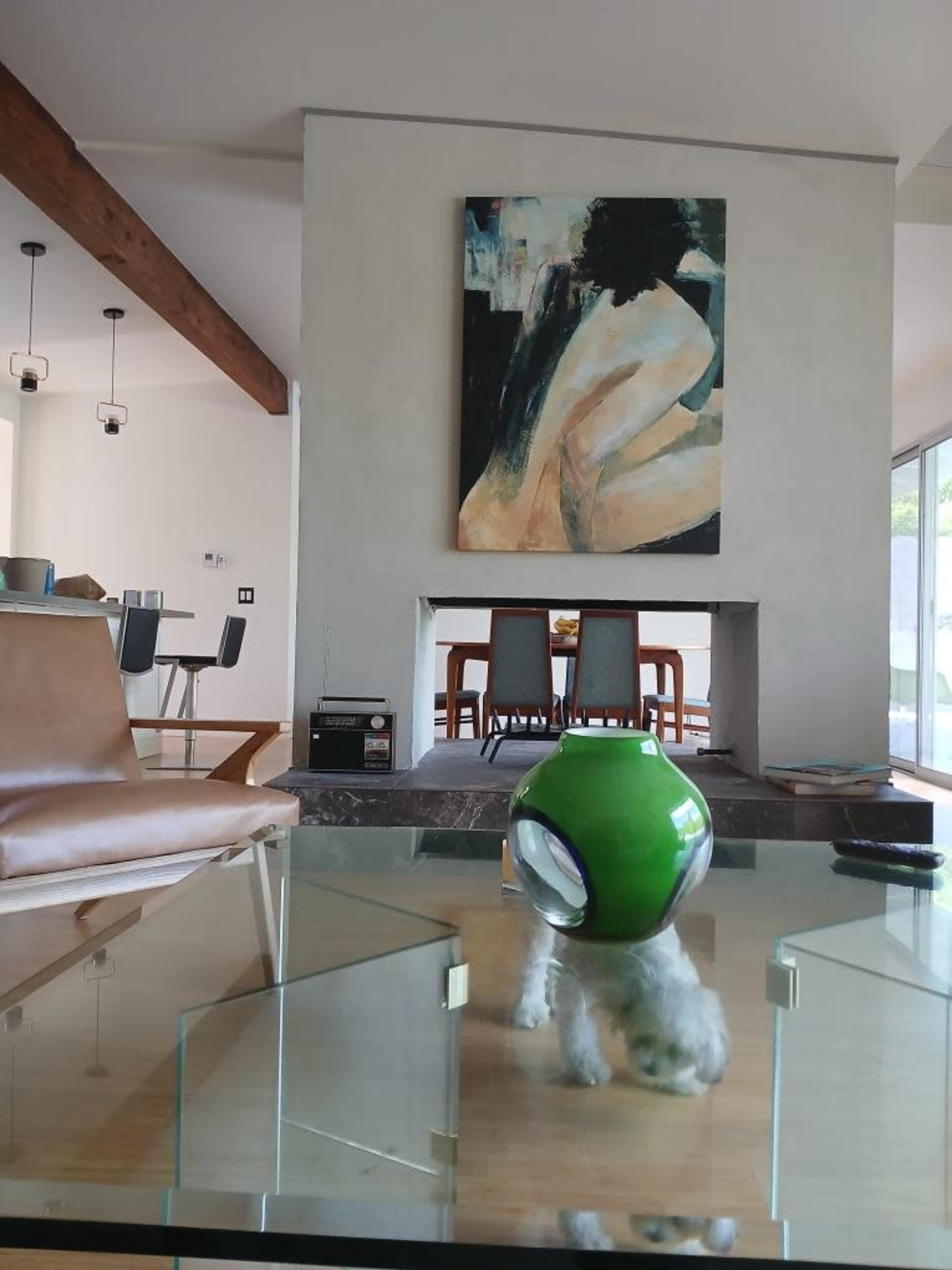 A modern living room features a glass coffee table with a green decorative bowl, a large abstract painting on the wall, and a dining area visible in the background.