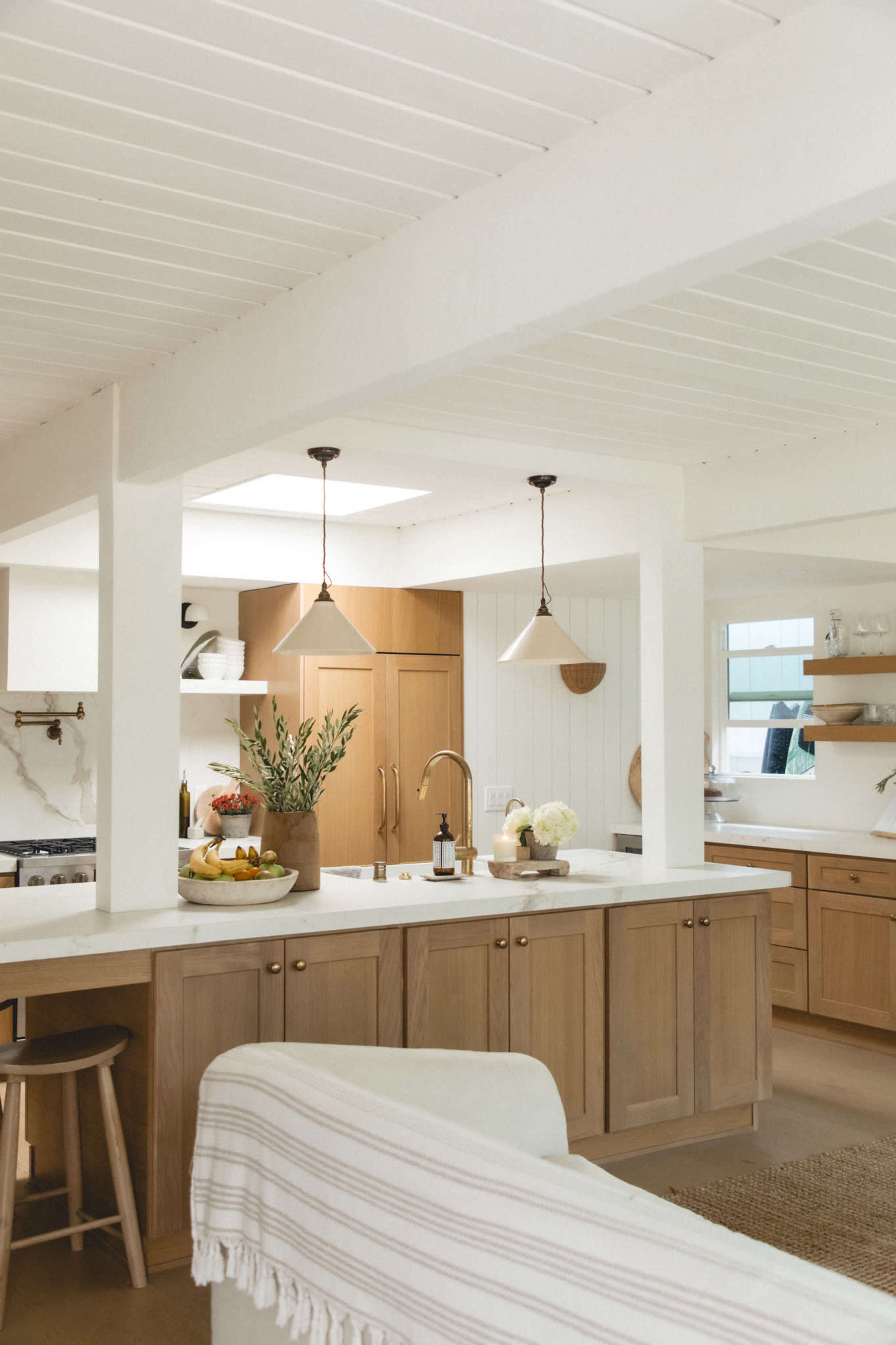 The image shows a bright, modern kitchen featuring wooden cabinets, a large island with a white countertop, and pendant lights hanging overhead.