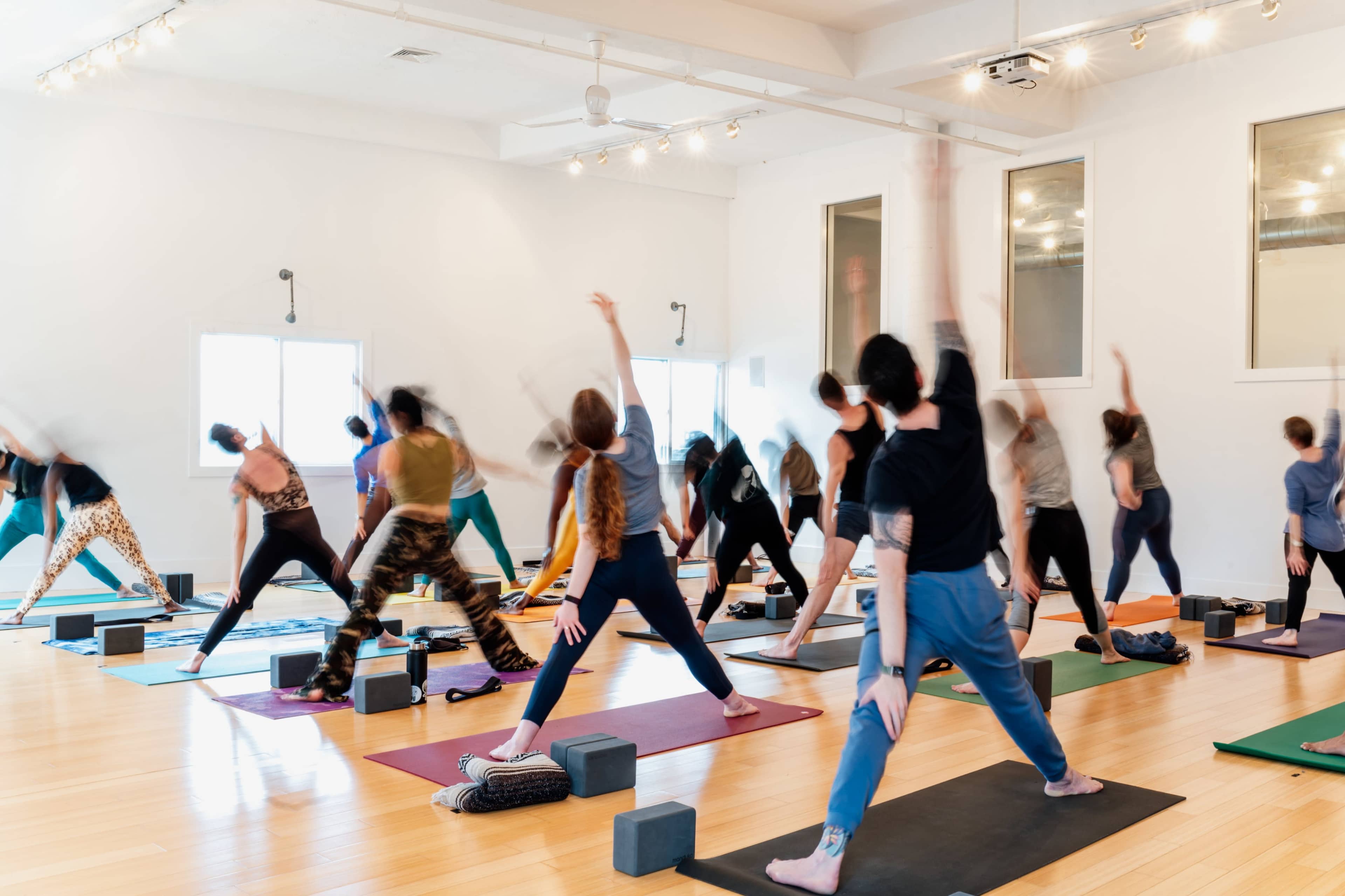 A group of people participates in a yoga class in a bright studio with mats and props arranged on a wooden floor.