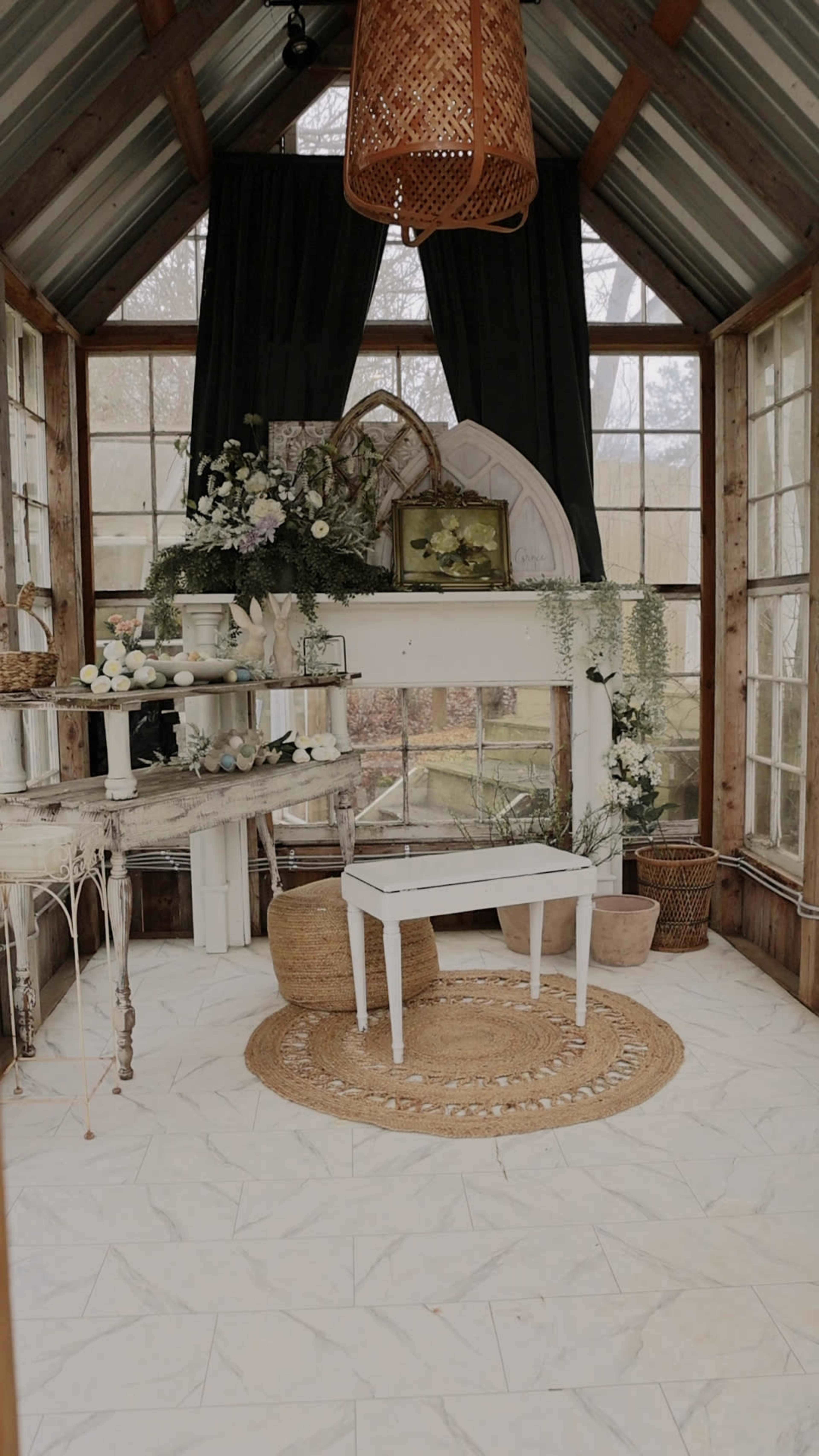 The image shows a cozy glass greenhouse with a white table, a round woven rug, and large windows filled with natural light.