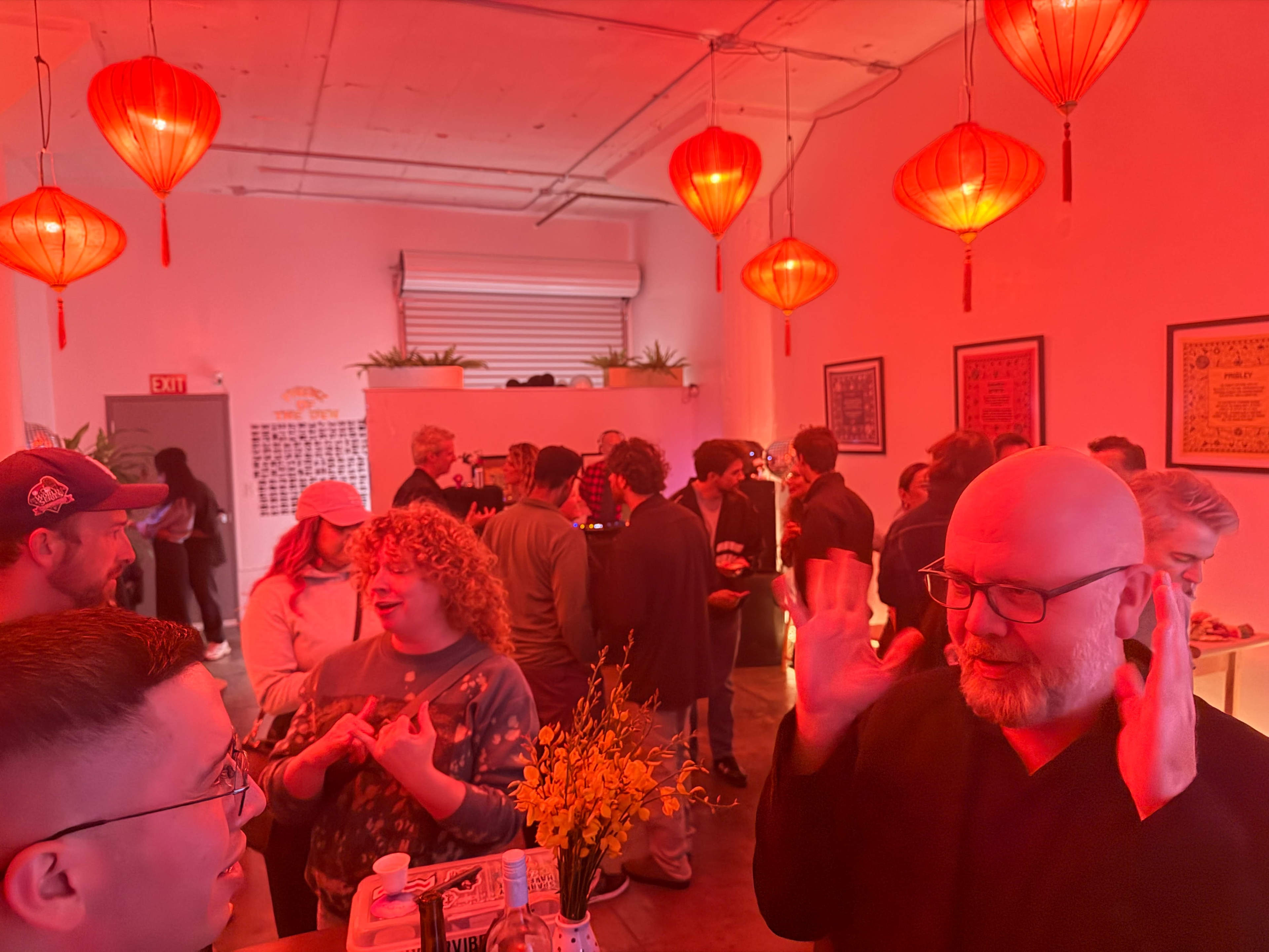 A crowd of people is gathered in a red-lit room decorated with hanging lanterns, engaging in conversation and socializing.