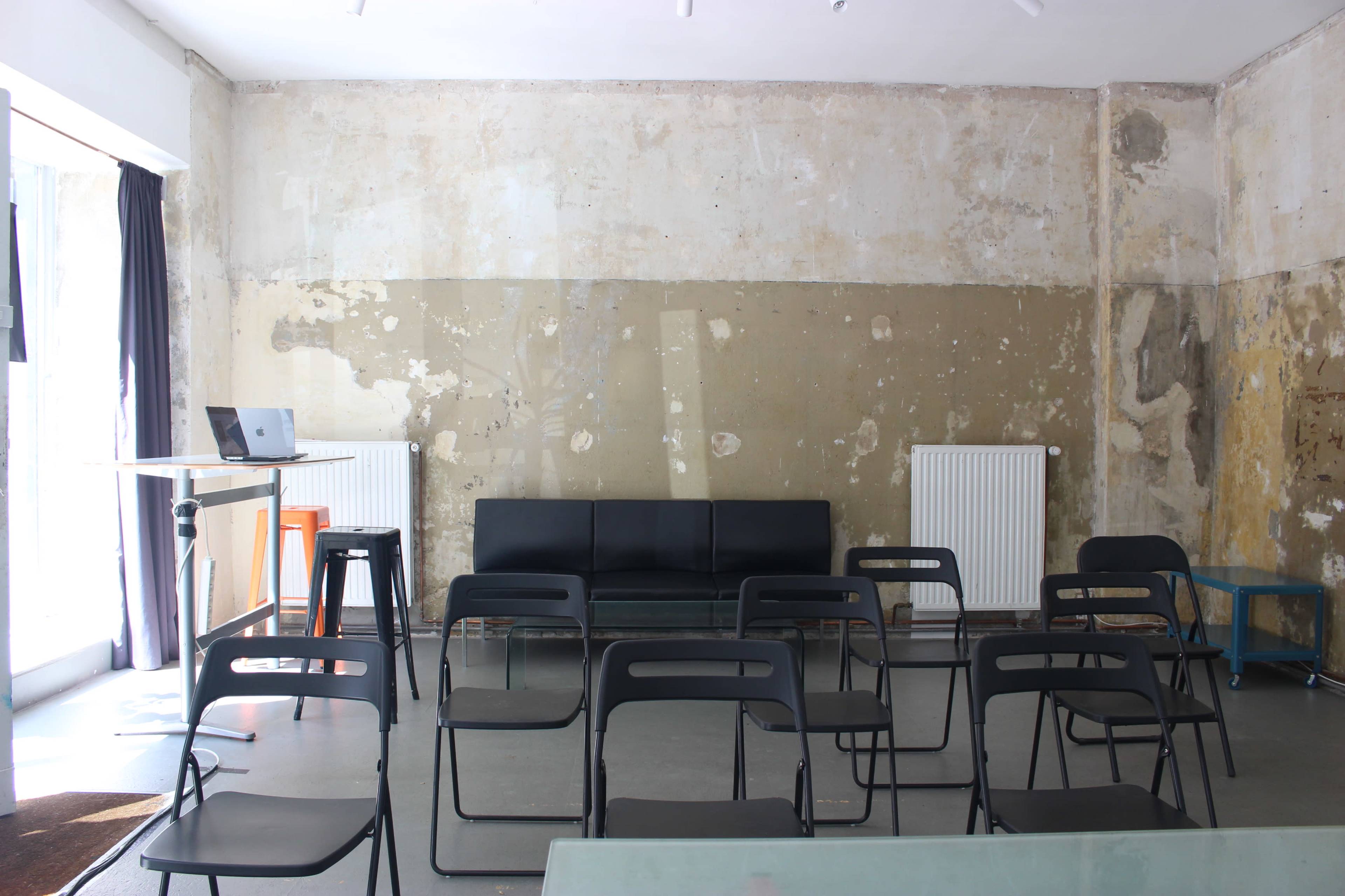 An empty room with a worn, partially peeling wall, containing a black sofa, scattered black chairs, a small table with a laptop, and a radiator.