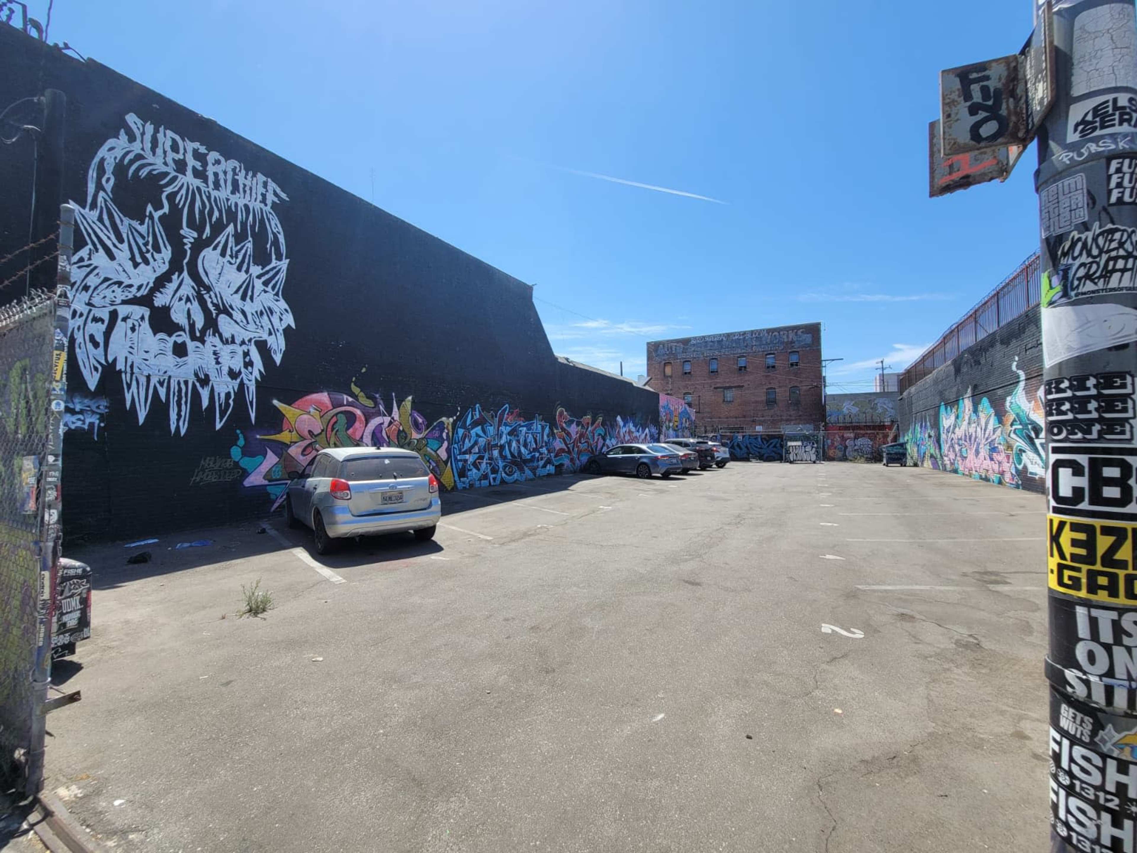 The image shows a vacant parking lot filled with graffiti-covered walls and a few parked cars.