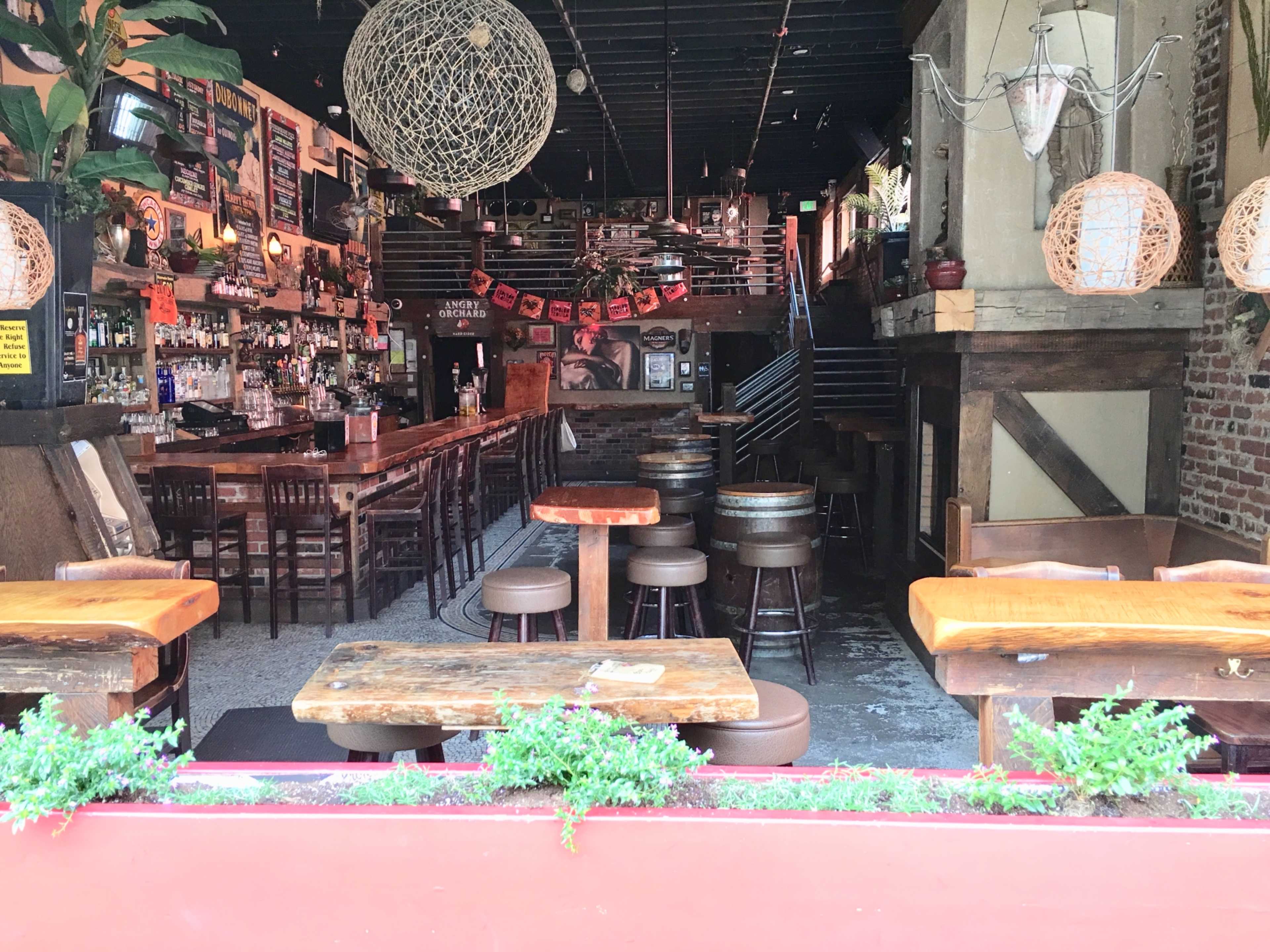 A rustic bar and dining area with wooden tables, a long bar counter, and decor including hanging light fixtures and plants.