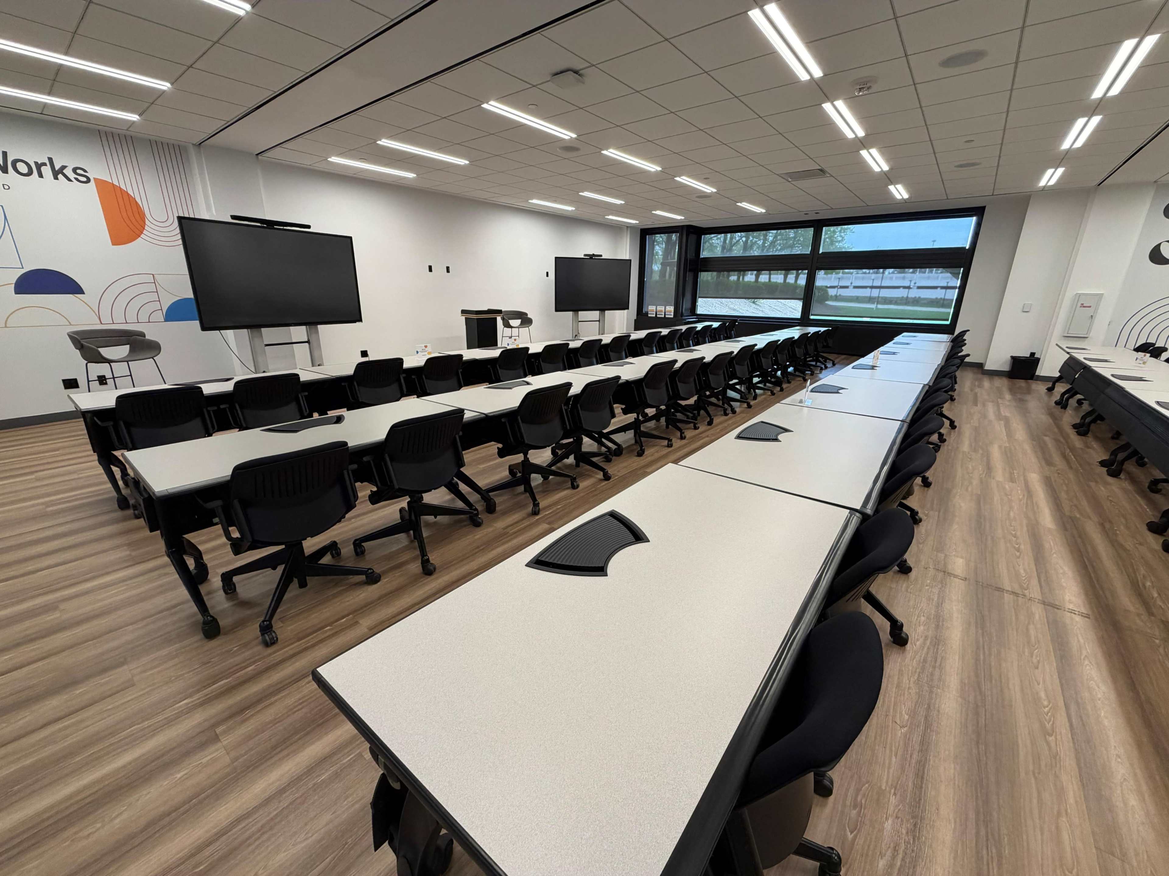 The image shows a modern classroom or conference room equipped with multiple rows of tables and chairs, large screens on the walls, and ample natural light from large windows.