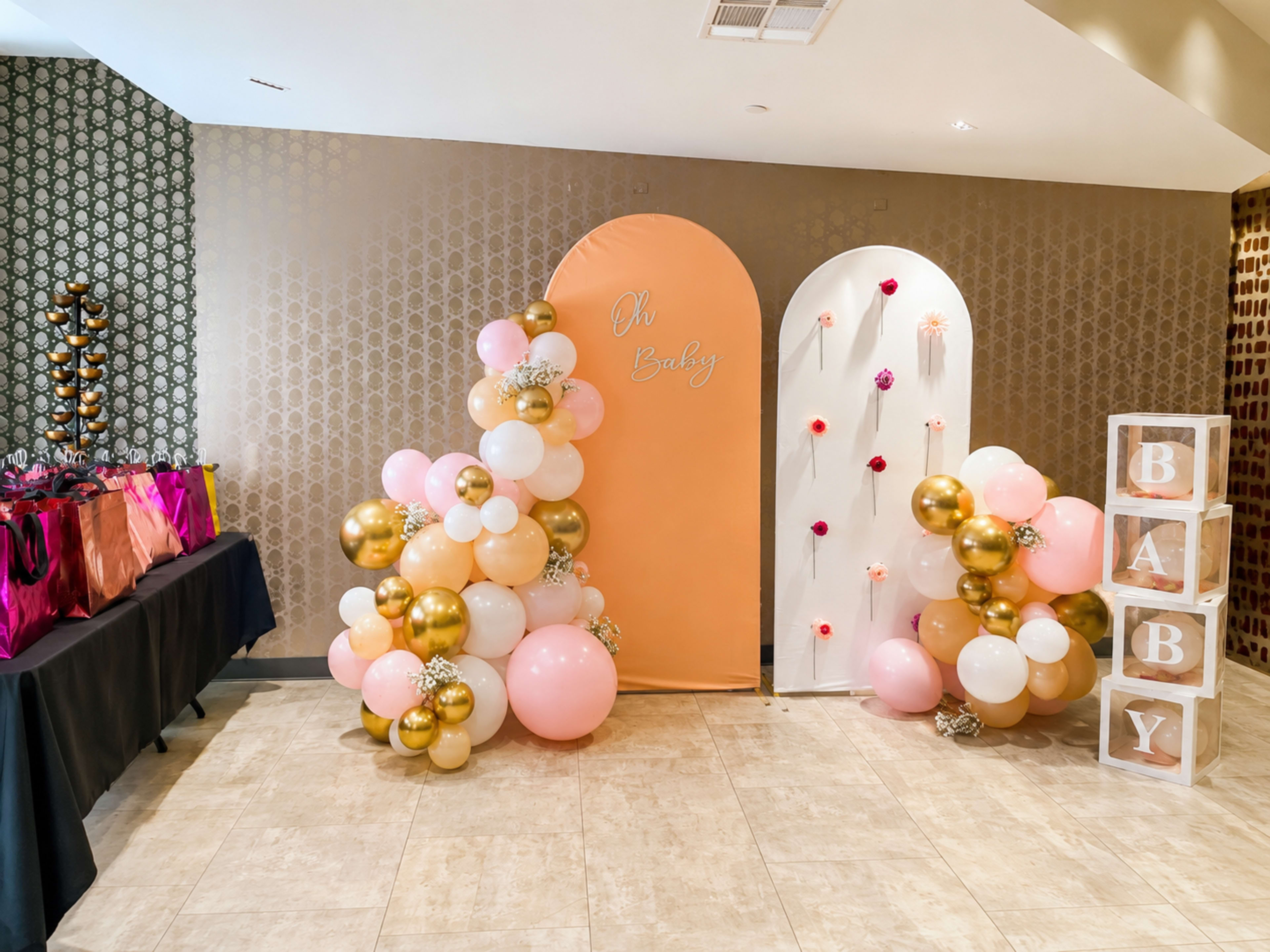 The image shows a decorated event space featuring two balloon arrangements, one with peach and gold balloons, and the other with white and pink balloons, alongside a decorative backdrop that reads "Oh Baby."