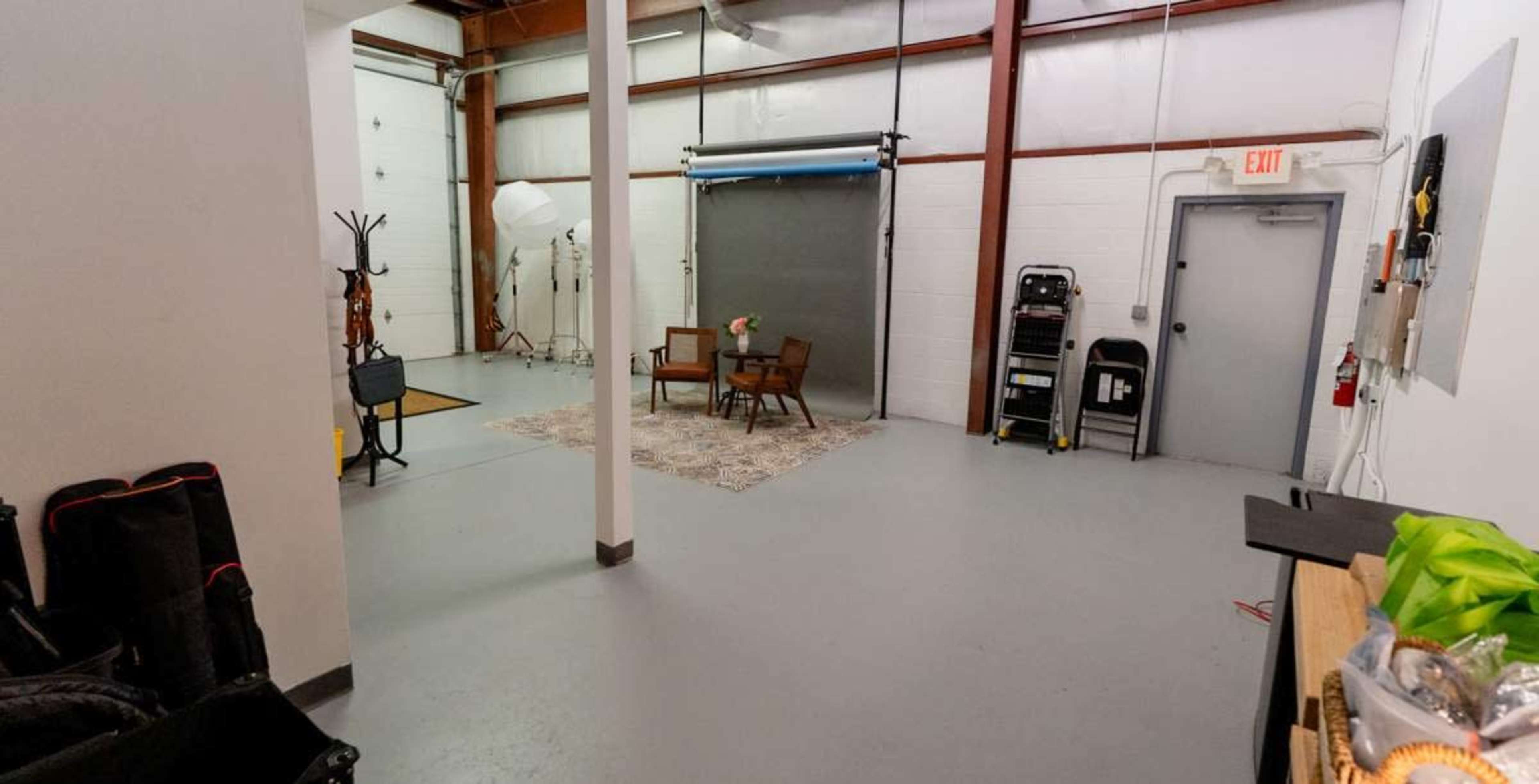 The image shows an industrial-style studio space with a rug and two chairs set up in the center, surrounded by photography equipment and a large backdrop.
