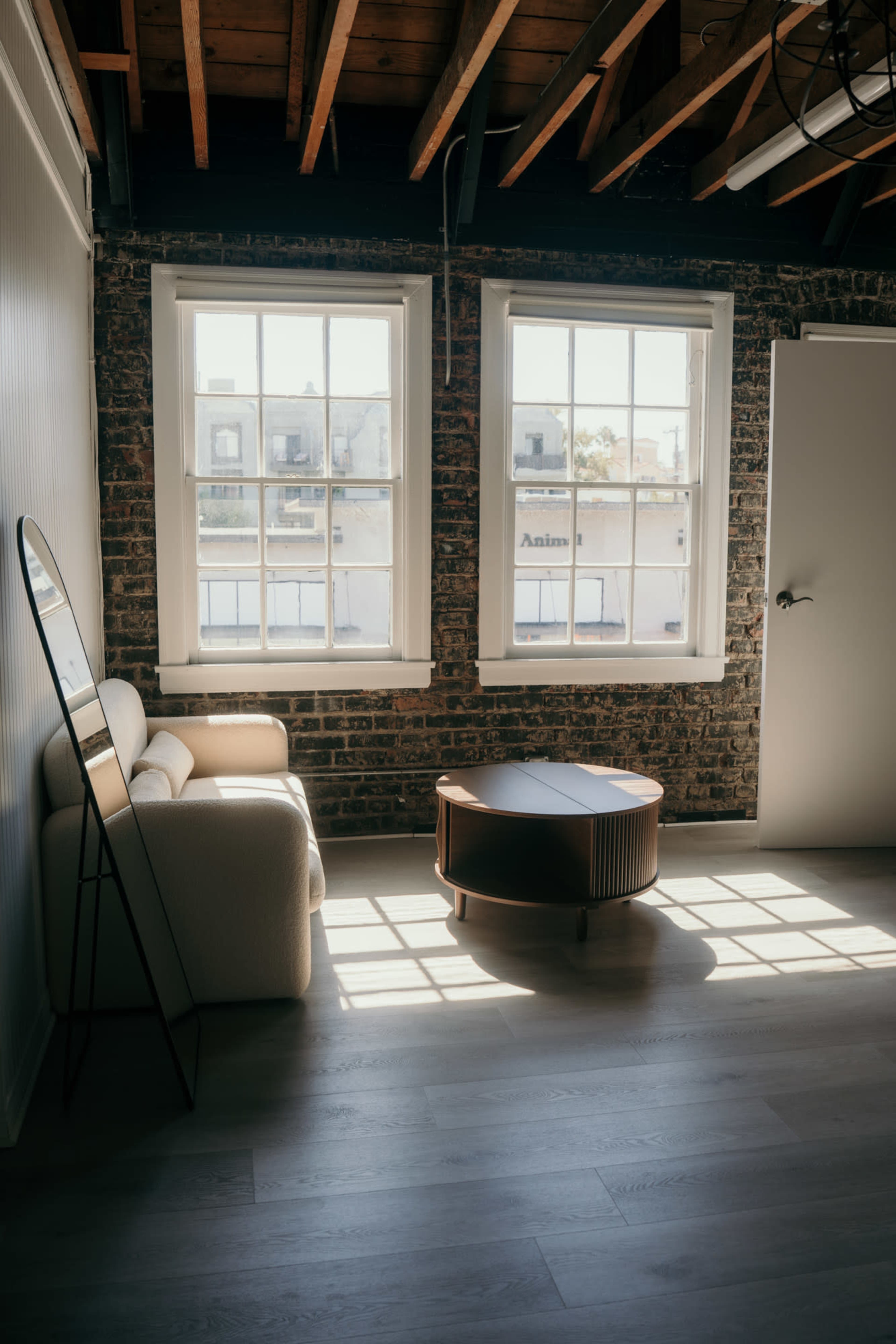The image shows a bright interior space with exposed brick walls, a light-colored sofa, a round wooden coffee table, and two large windows allowing sunlight to stream in.