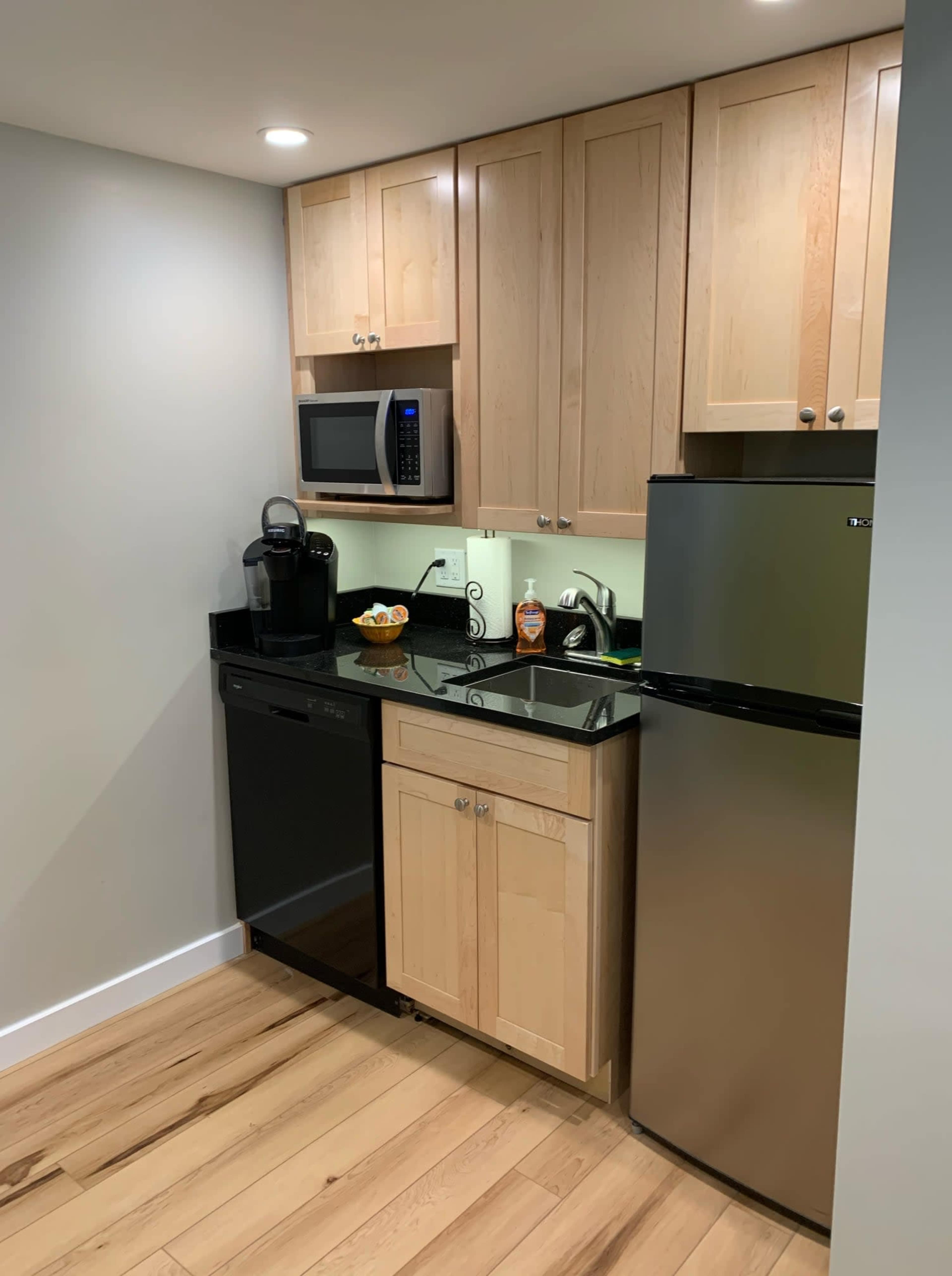 The image shows a kitchen with wooden cabinets, a black countertop, a microwave, a coffee maker, and a black dishwasher and refrigerator.