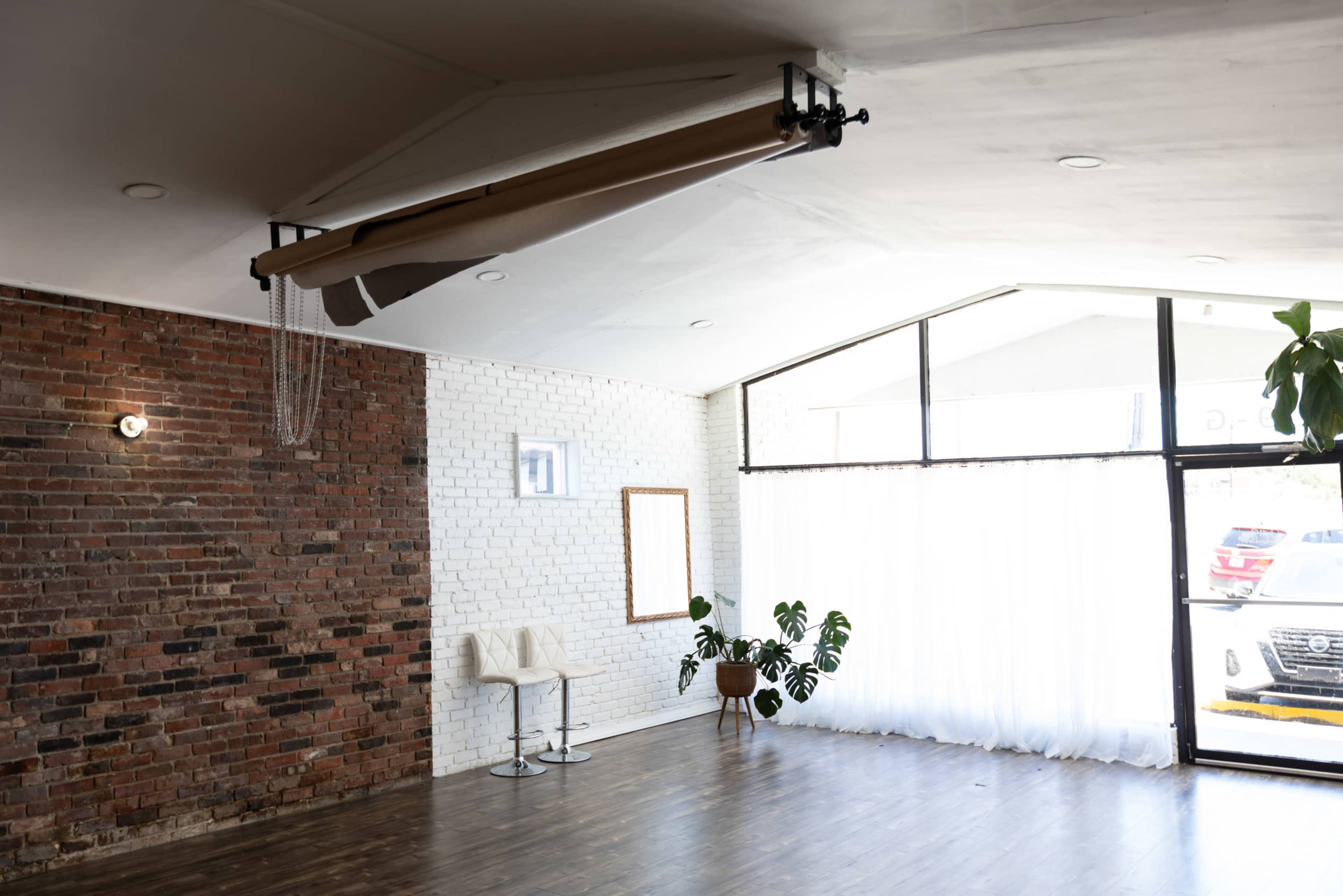 The image shows a modern, sparsely furnished studio with exposed brick walls, large windows, and a few indoor plants.