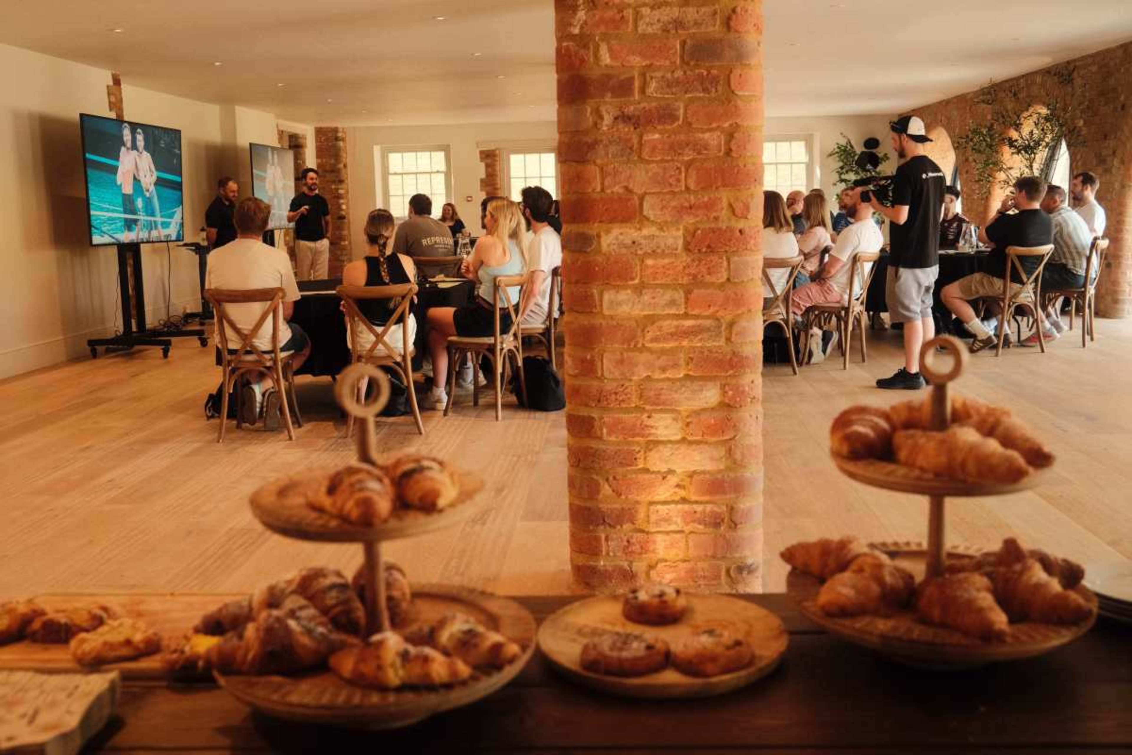 The image shows a group of people seated at a table in a well-lit room, while an audience watches a presentation on a screen, with a table of pastries in the foreground.