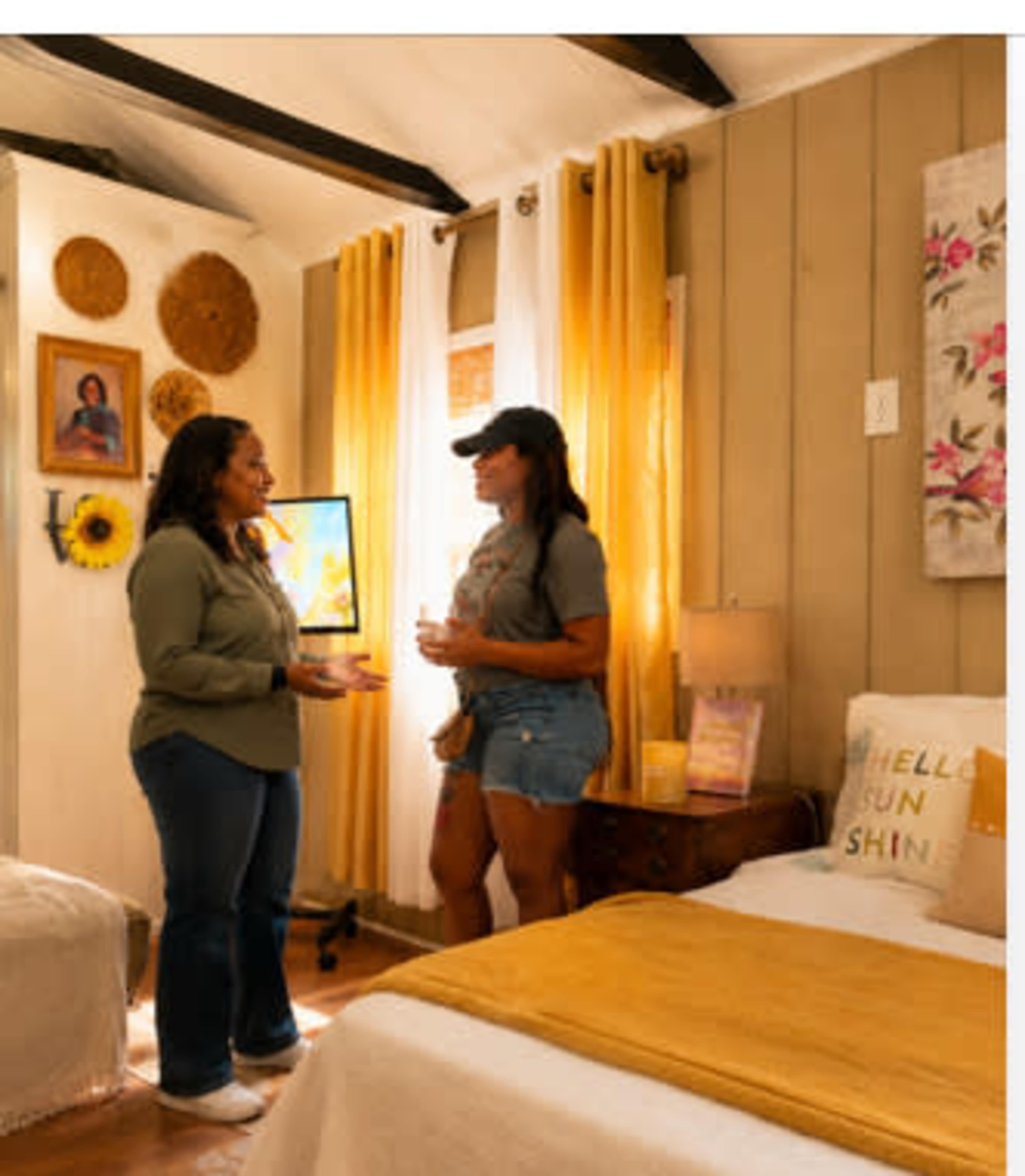 Two women are standing in a warmly decorated bedroom featuring yellow curtains, floral decor, and two beds.