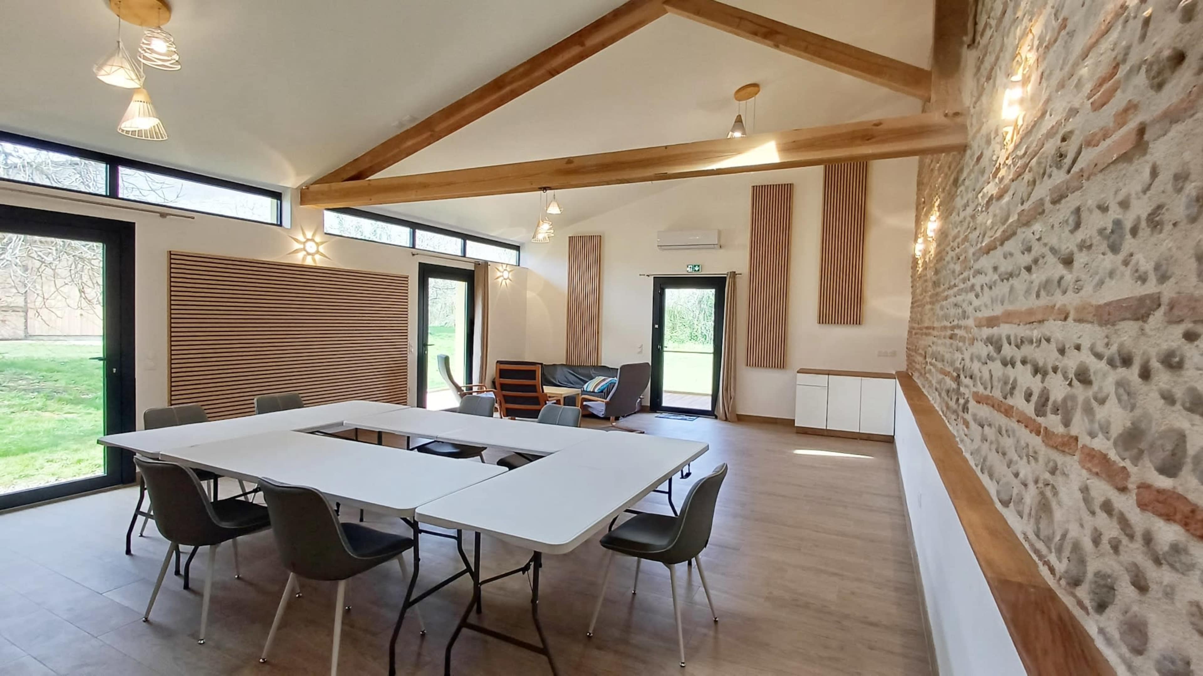 The image shows a modern meeting room with a large white table surrounded by chairs, large windows providing natural light, and stone and wood accents on the walls.