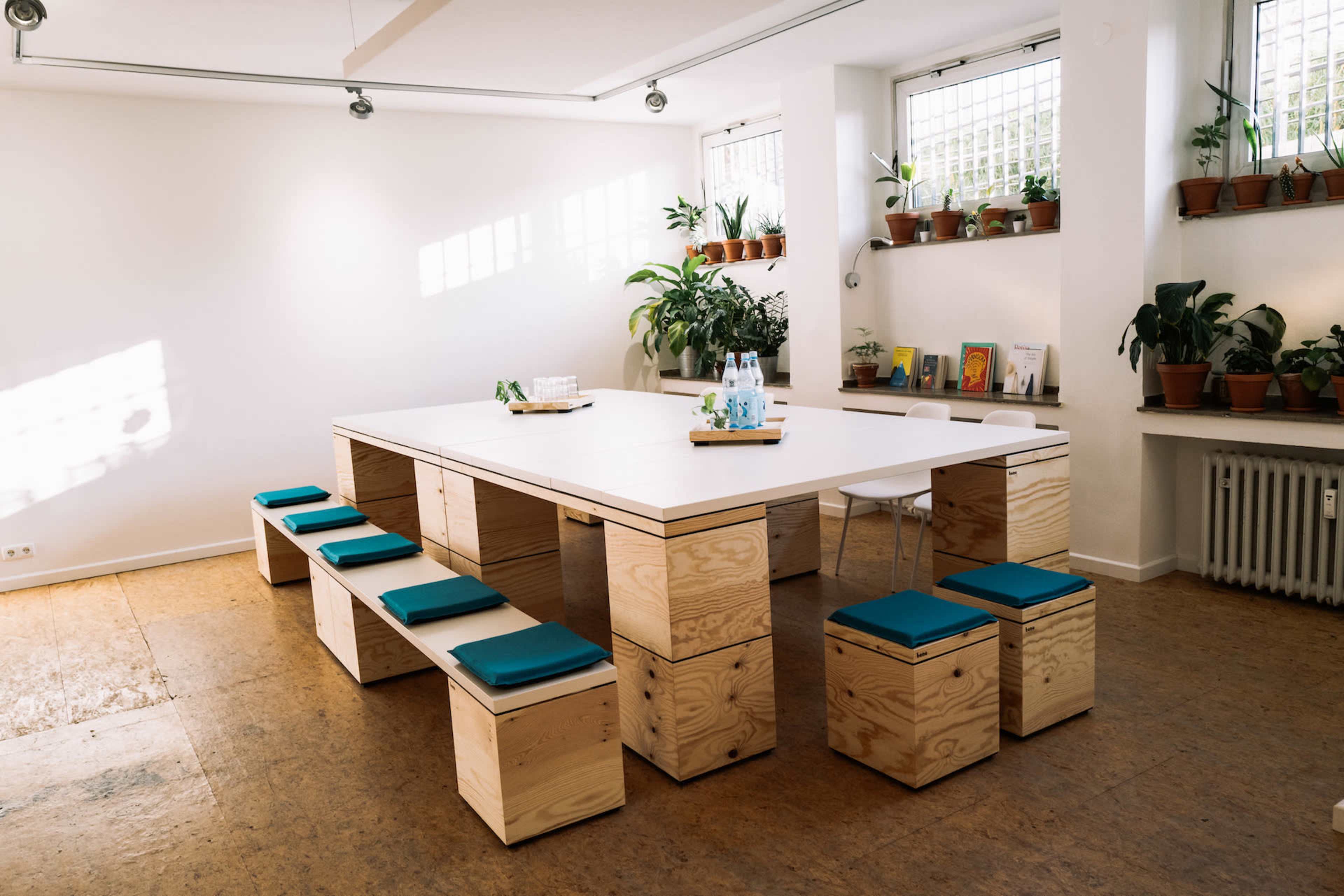 A spacious, well-lit meeting room features a large white table surrounded by wooden benches with teal cushions and potted plants lining the windowsill.