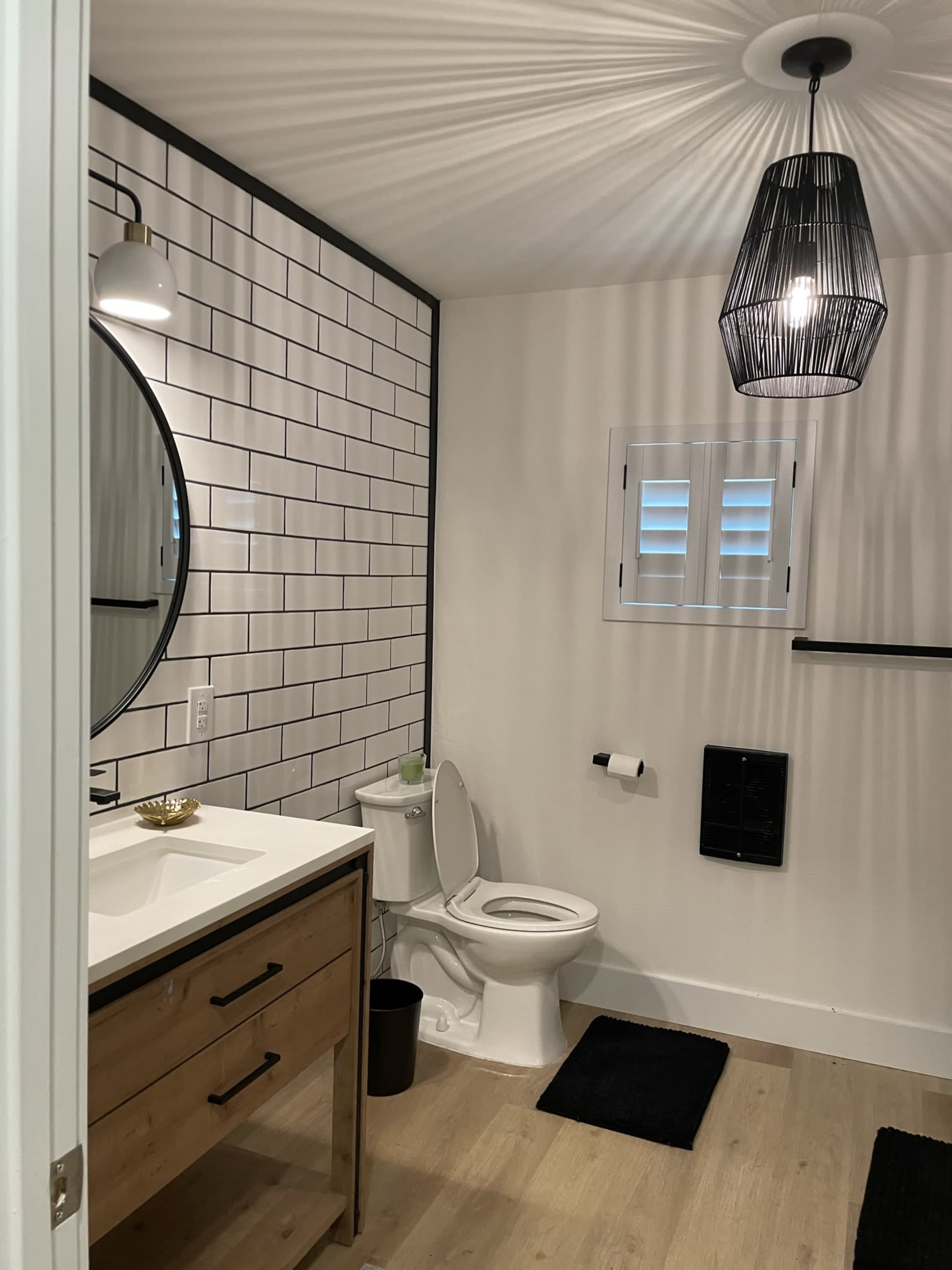 A bathroom with a round mirror above a wooden vanity, a white toilet, and black accent features against a white subway tile wall.