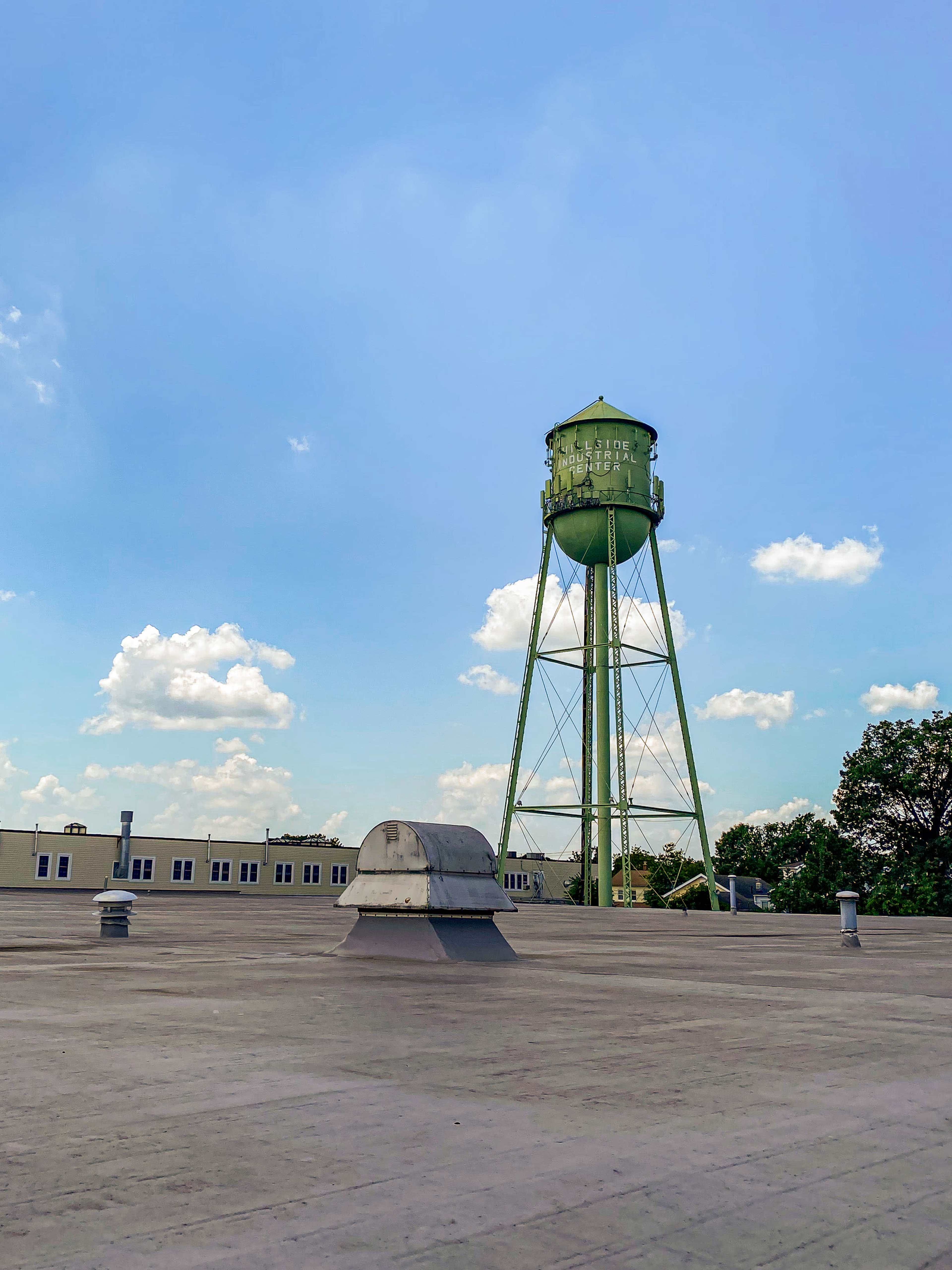 A green water tower stands tall against a blue sky, with scattered clouds and a flat rooftop in the foreground.