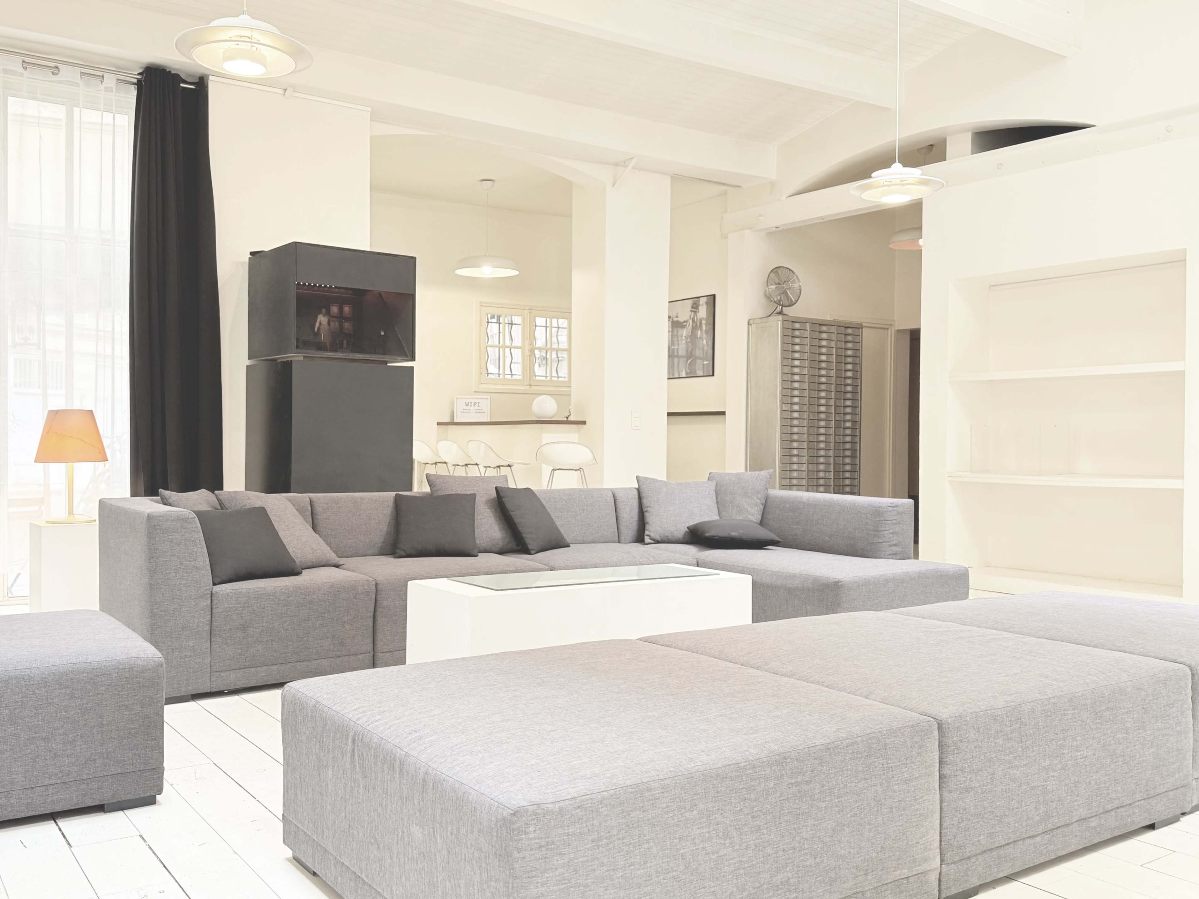 The image shows a modern living room with a large gray sectional sofa, a glass coffee table, and minimalistic decor against white walls and wooden floors.