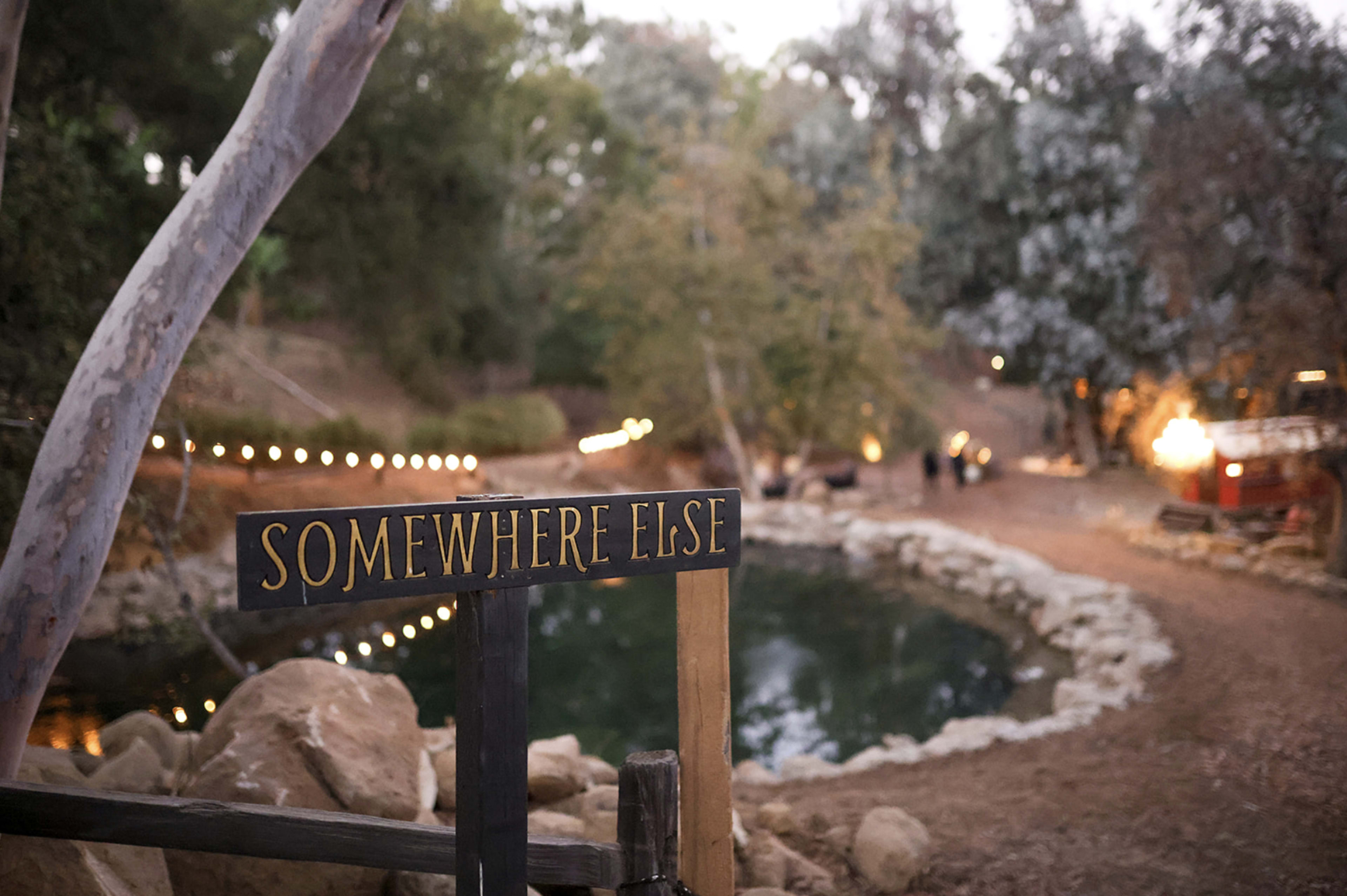 A wooden sign reading "SOMEWHERE ELSE" stands near a tranquil pond surrounded by trees and soft evening lights.